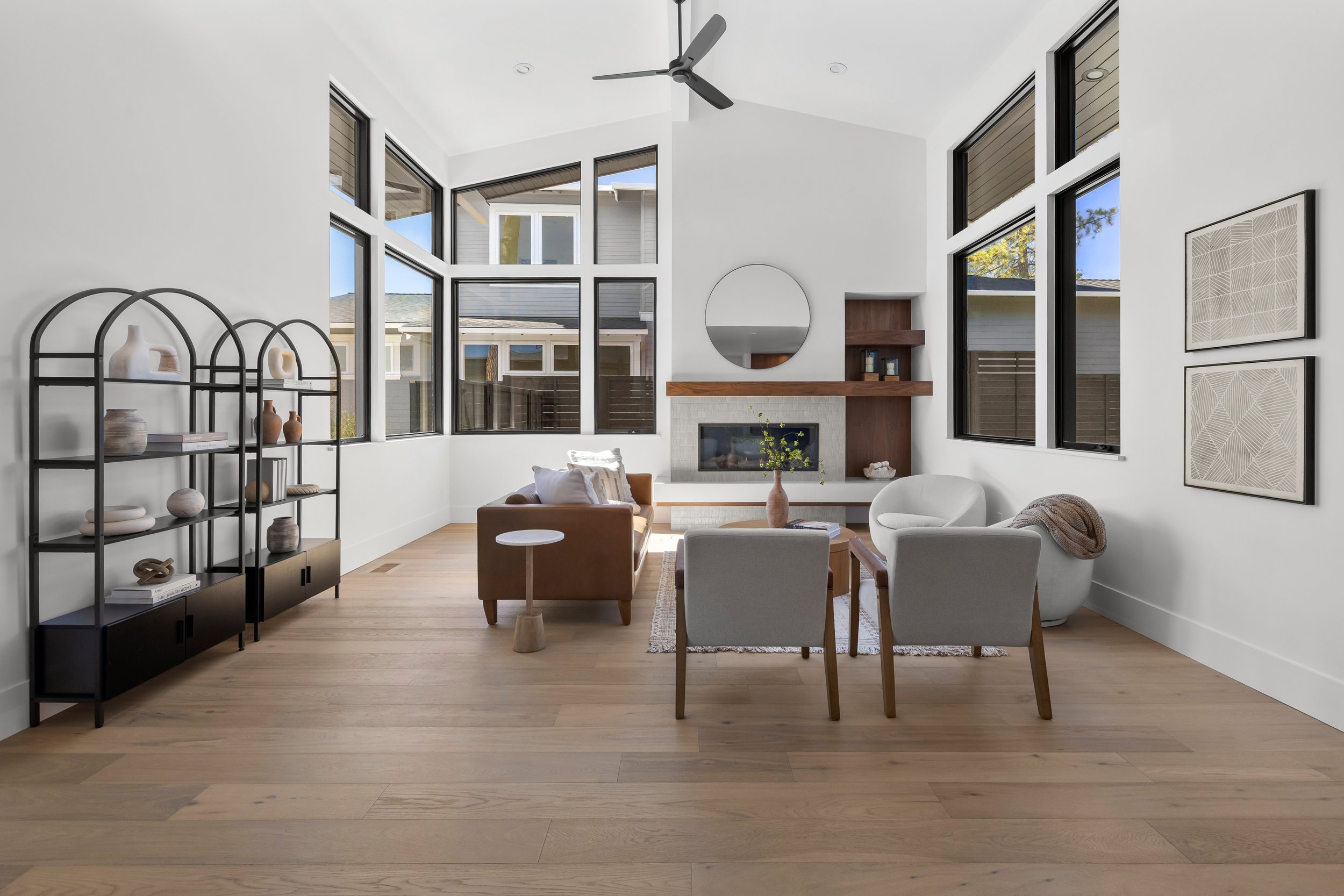 Modern living room with large windows, white walls, wooden flooring, a fireplace with wooden mantel, and contemporary furniture including a white armchair, beige chair, and brown sofa. There's a black metal shelving unit with vases and decor, and geometric artwork on the wall.