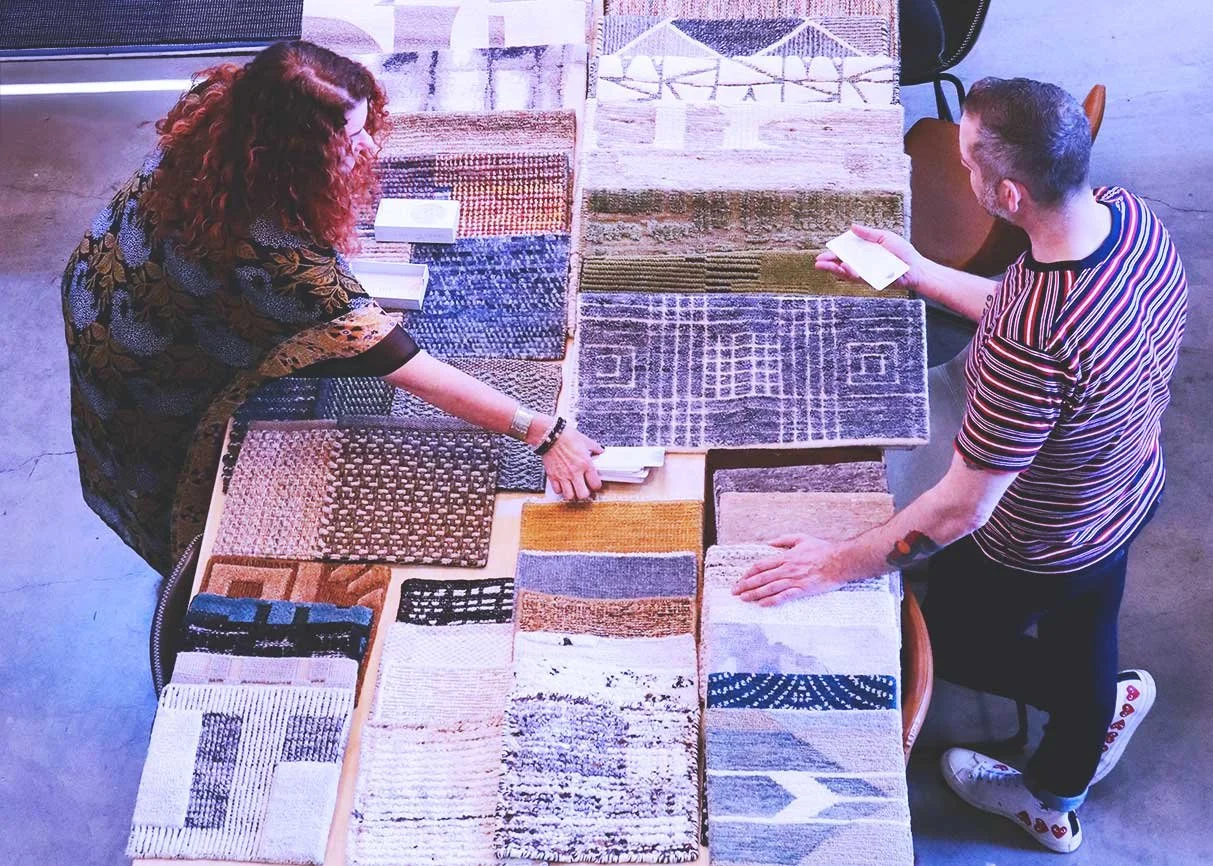Two people shopping for rugs at a store, looking at a variety of patterned rugs laid out on a table.