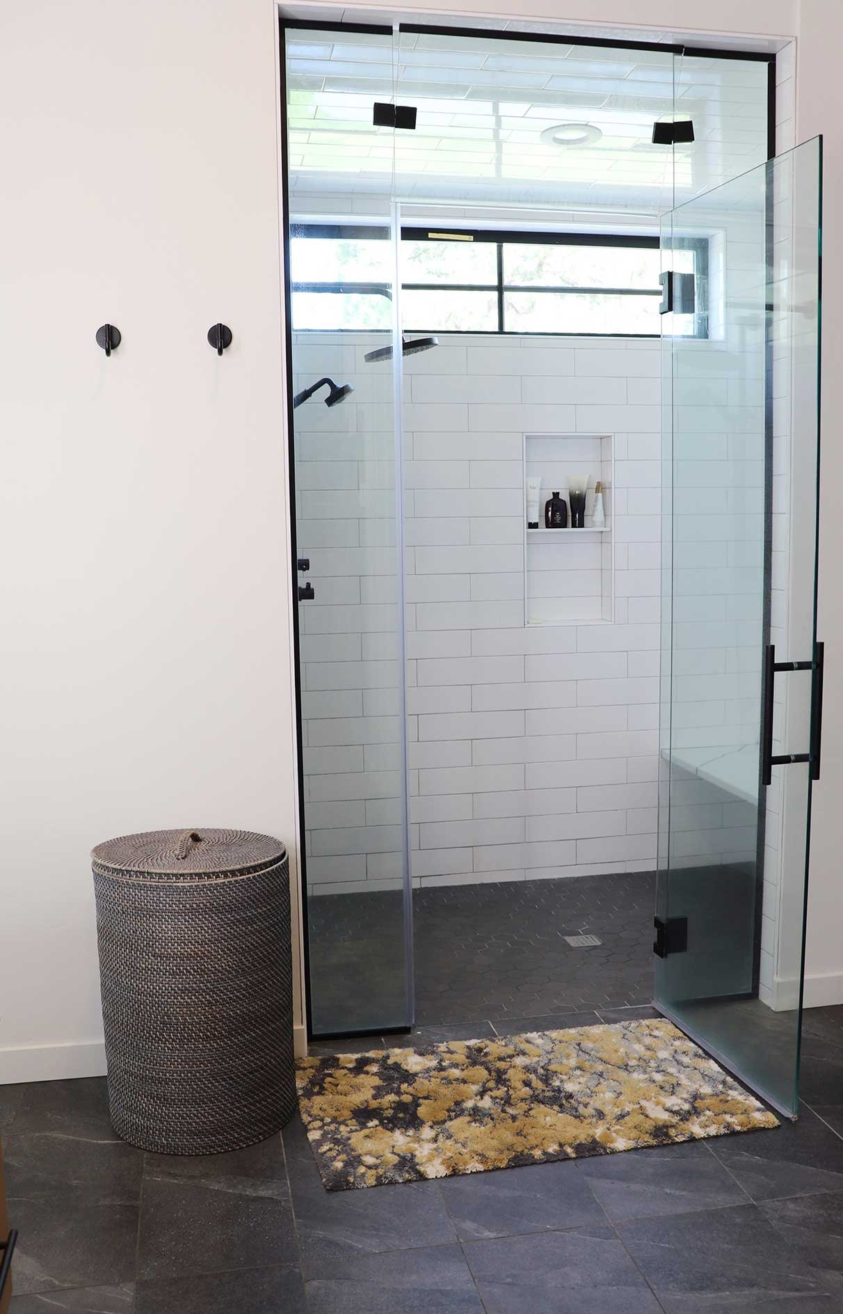 Modern bathroom with a glass walk-in shower, black fixtures, a white tiled wall, small niche with toiletries, a patterned round laundry basket, and a floral bathroom rug on dark tile floor.