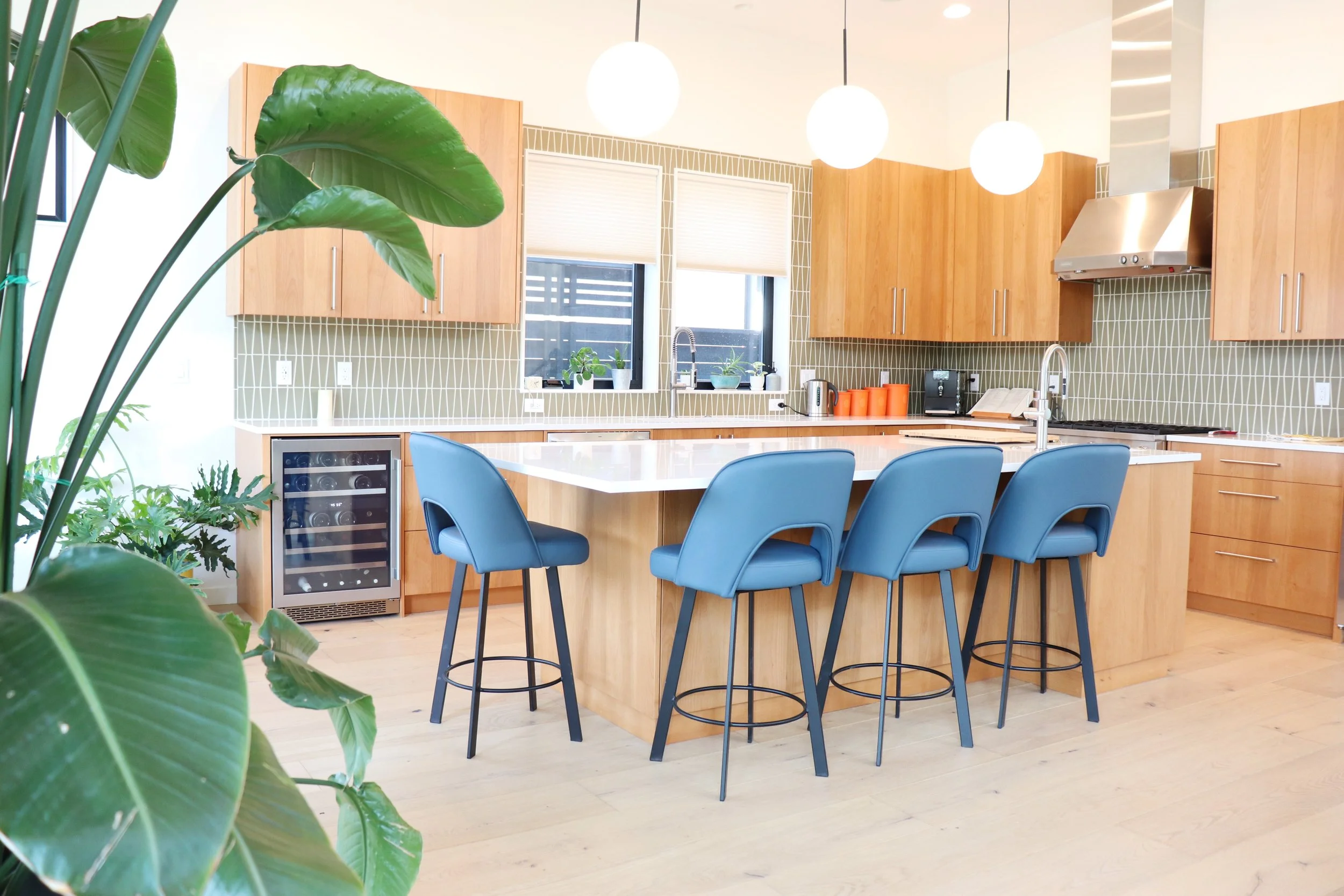 Modern kitchen design with light wood cabinets, a white countertop island with four blue chairs, green plants near windows, and hanging round white lights.
