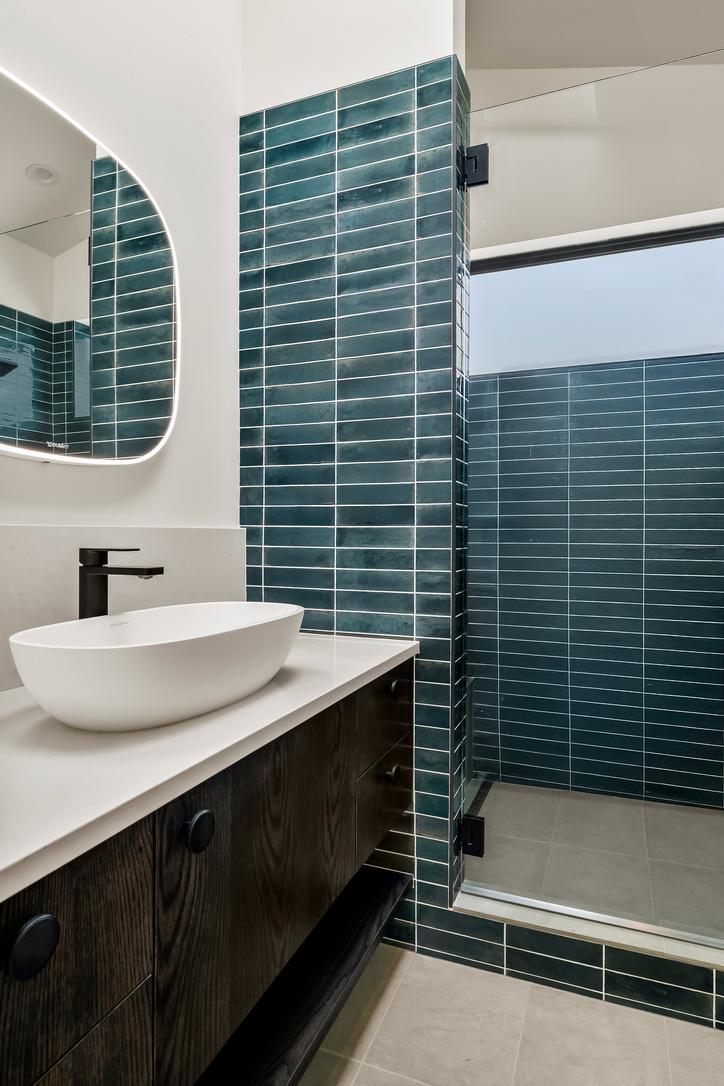 Modern bathroom interior design featuring a vessel sink on a white countertop, dark wooden vanity, and a walk-in shower with teal tiles and a glass door.