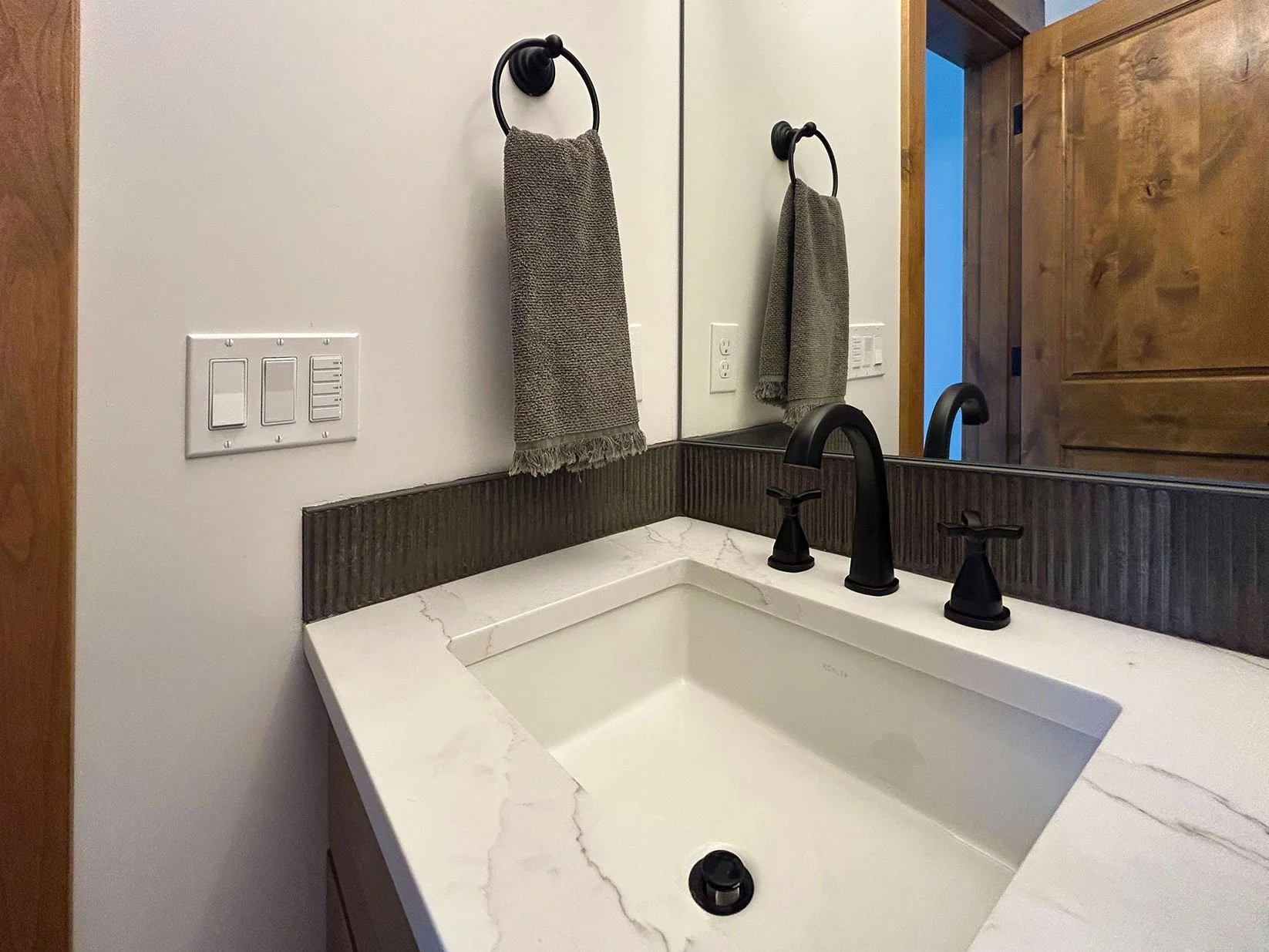 Bathroom vanity with a white marble countertop, black faucet, and a large mirror in Bend, Oregon. Two gray towels hang on black ring towel holders. Wooden door and wall are visible.