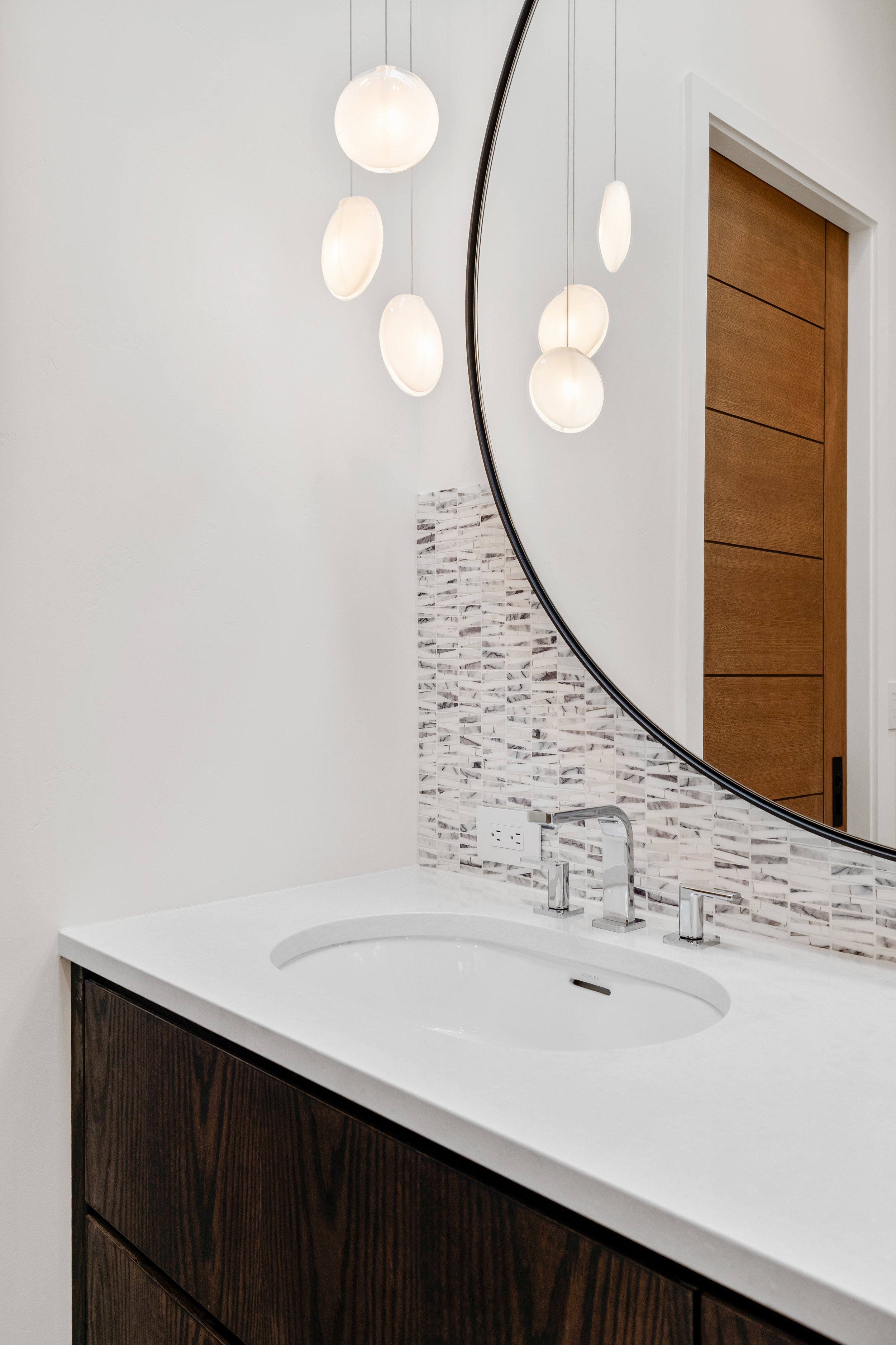 A modern bathroom vanity with white countertop, white sink, chrome faucet, large round mirror, textured mosaic tile backsplash, and a wooden door visible in the reflection.