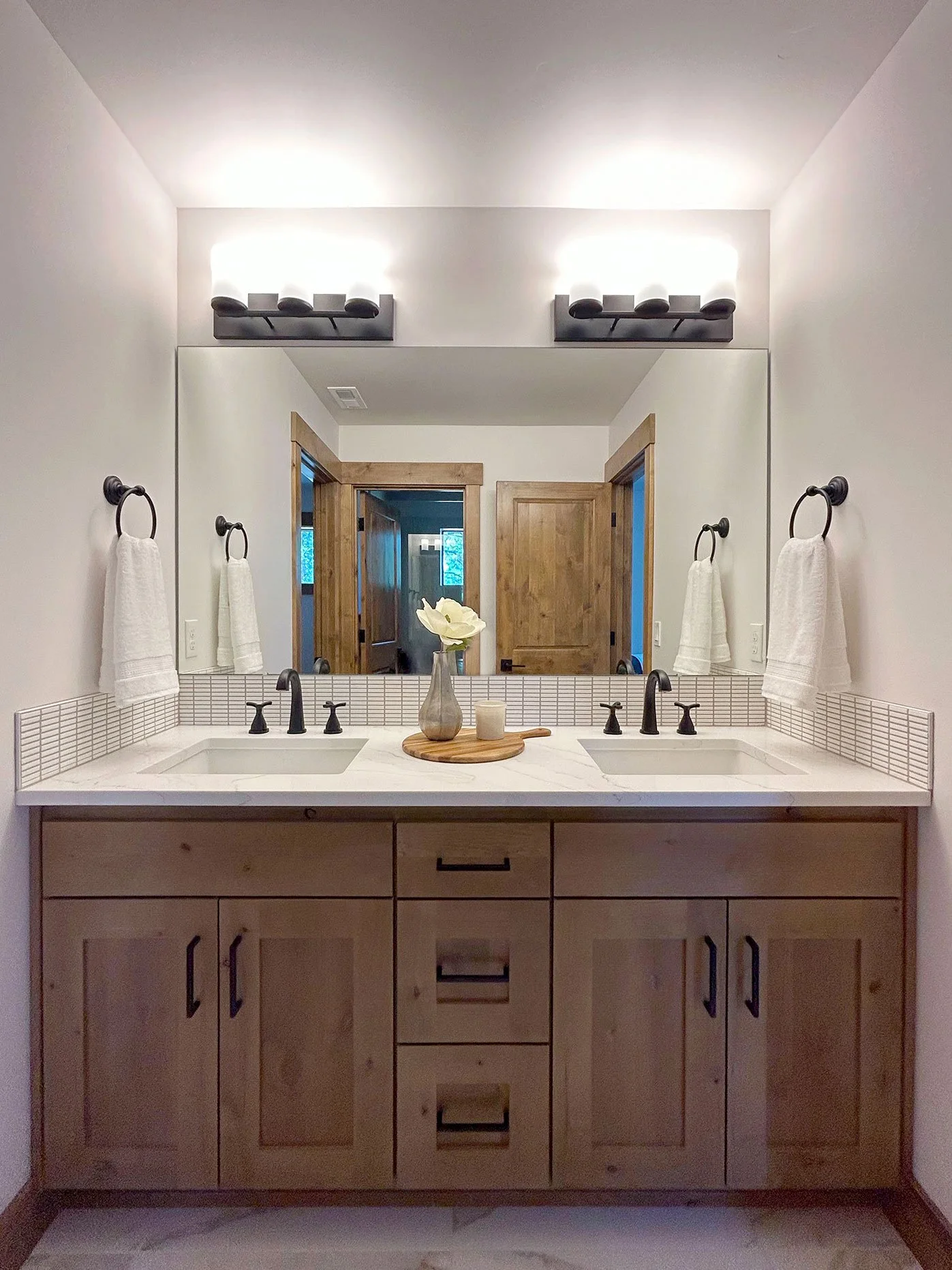 Bathroom double vanity with wooden cabinet, white countertop, black fixtures, large mirror, wall-mounted lights, and three white towels on black rings in Bend, Oregon.