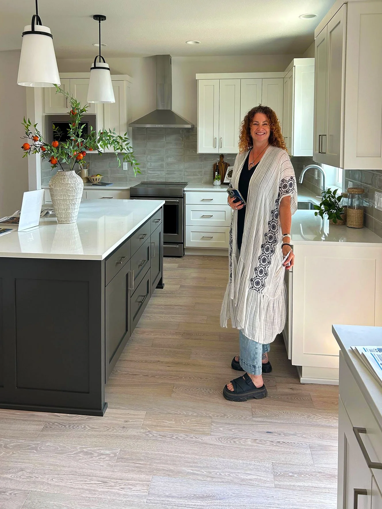 An interior designer standing in a modern kitchen smiling, wearing a long patterned white coat, black top, blue jeans, and black sandals in Bend, Oregon.