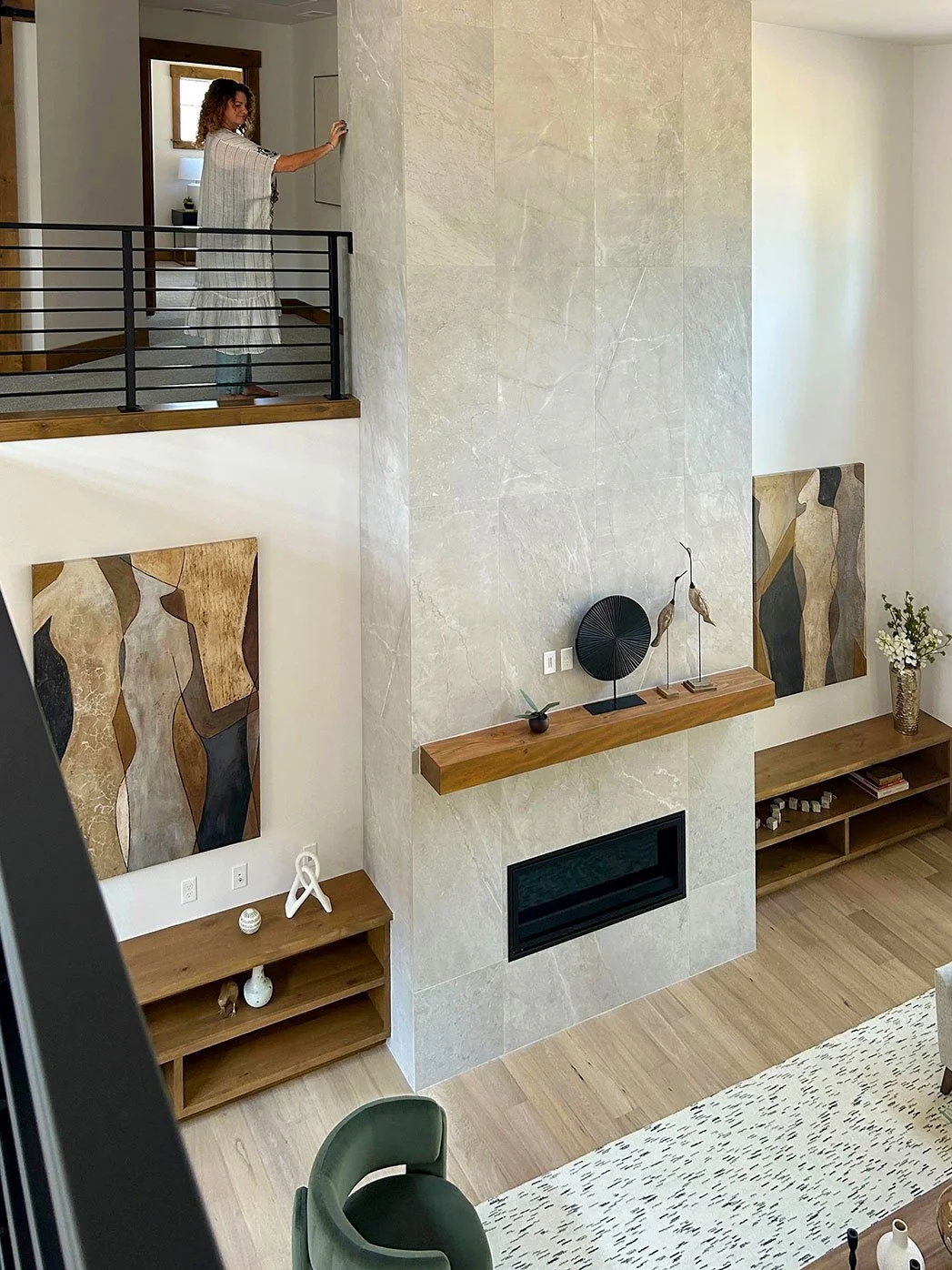 A woman in a striped dress stands on a second-level balcony of a modern living room, touching a wall. The living room features a large beige marble fireplace with a wooden mantel, abstract artwork on the walls, and minimalistic decorative sculptures