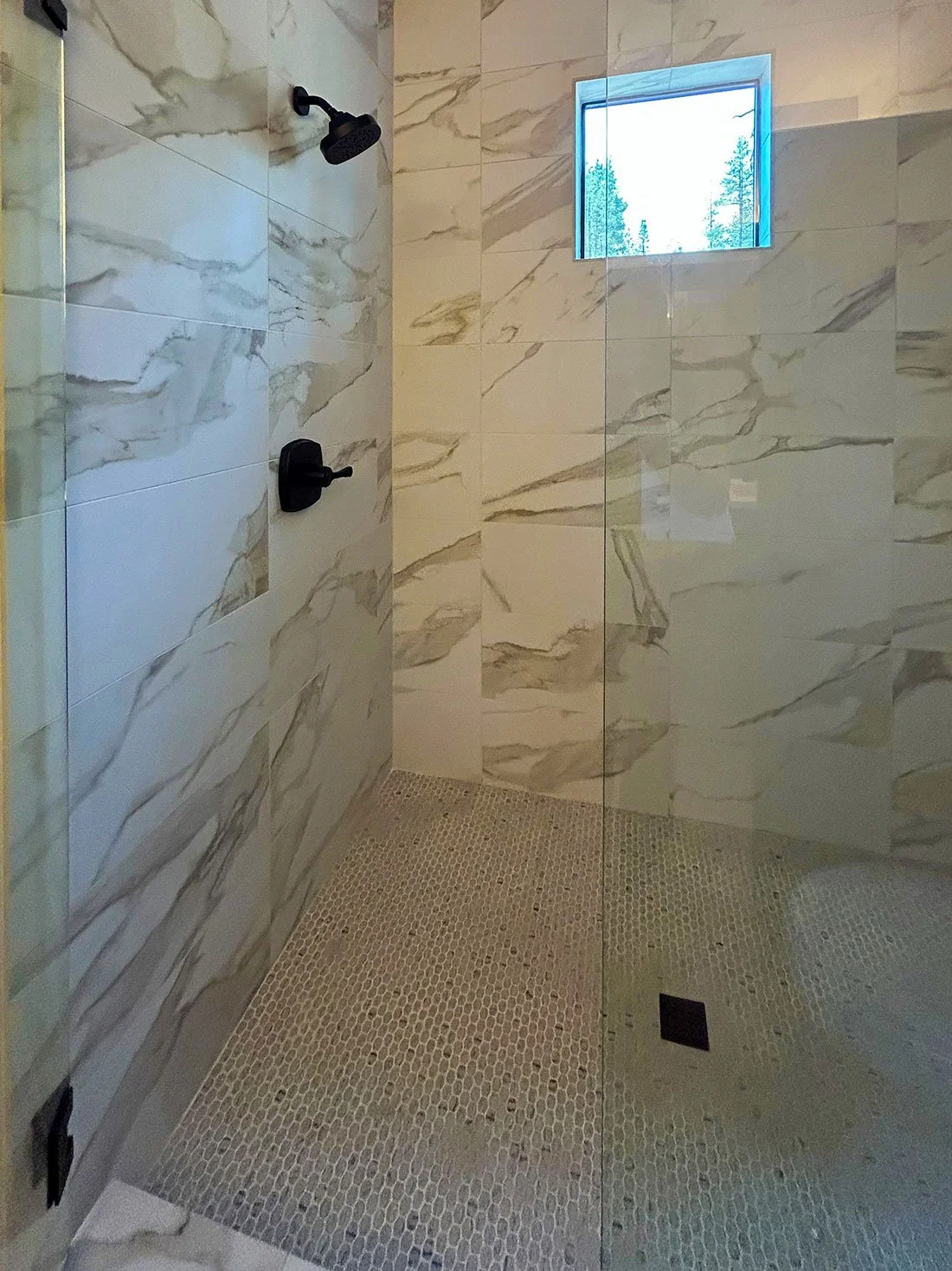 A modern walk-in shower with white marble-like tiles, black fixtures, a small rectangular window, and a pebble stone floor in Bend, Oregon.
