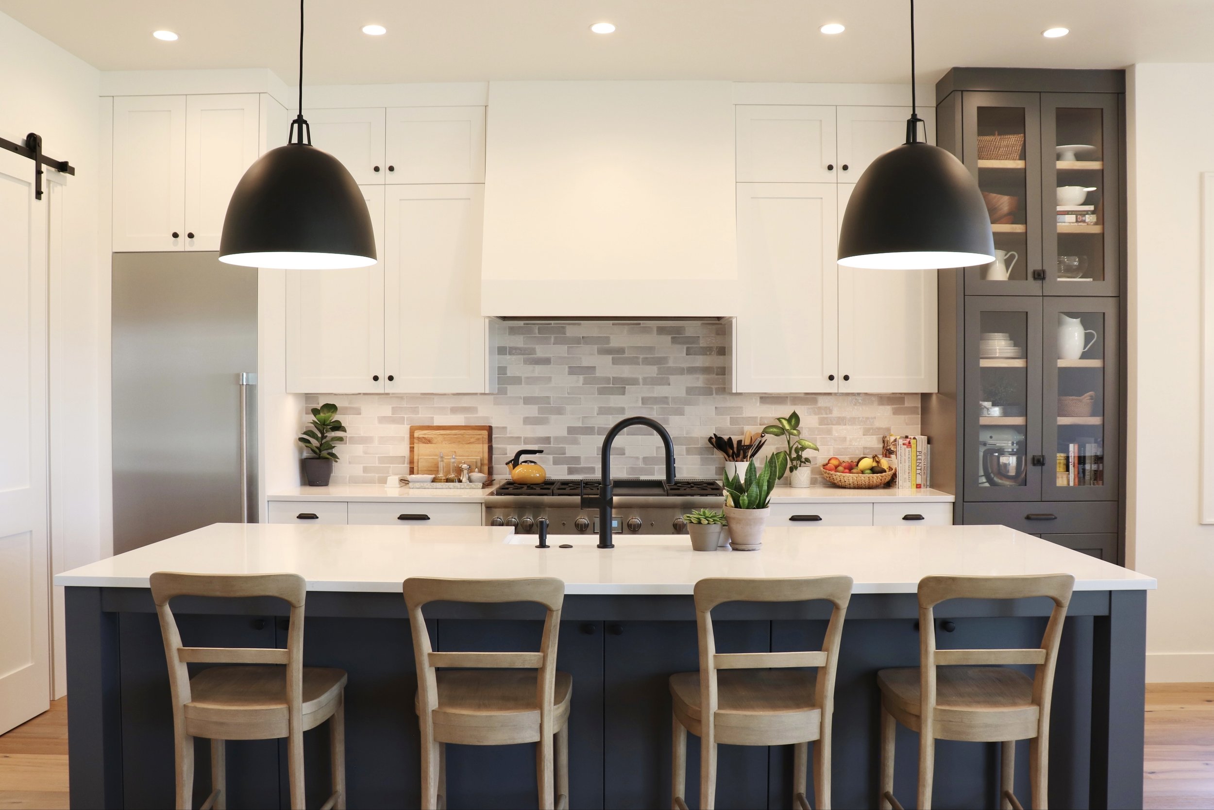 Modern kitchen in Bend, Oregon with white cabinets, dark gray island, four wooden chairs, black pendant lights, gray backsplash, and kitchenware.
