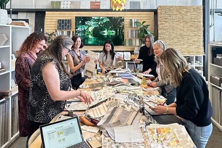 Group of women in a room looking at fabric samples and patterns on a long table, with a television, shelves, and a computer in the background.