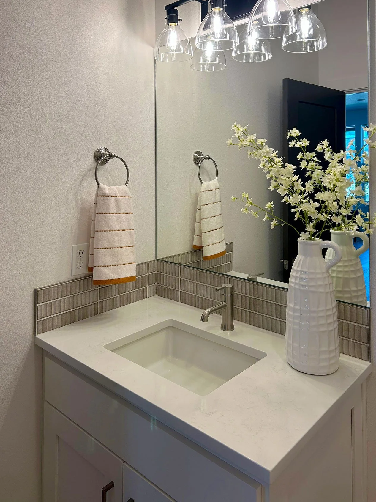 Bathroom vanity with a rectangular mirror above, a faucet, beige hand towels, a white vase with white flowers, and ceiling lights in Bend, Oregon.
