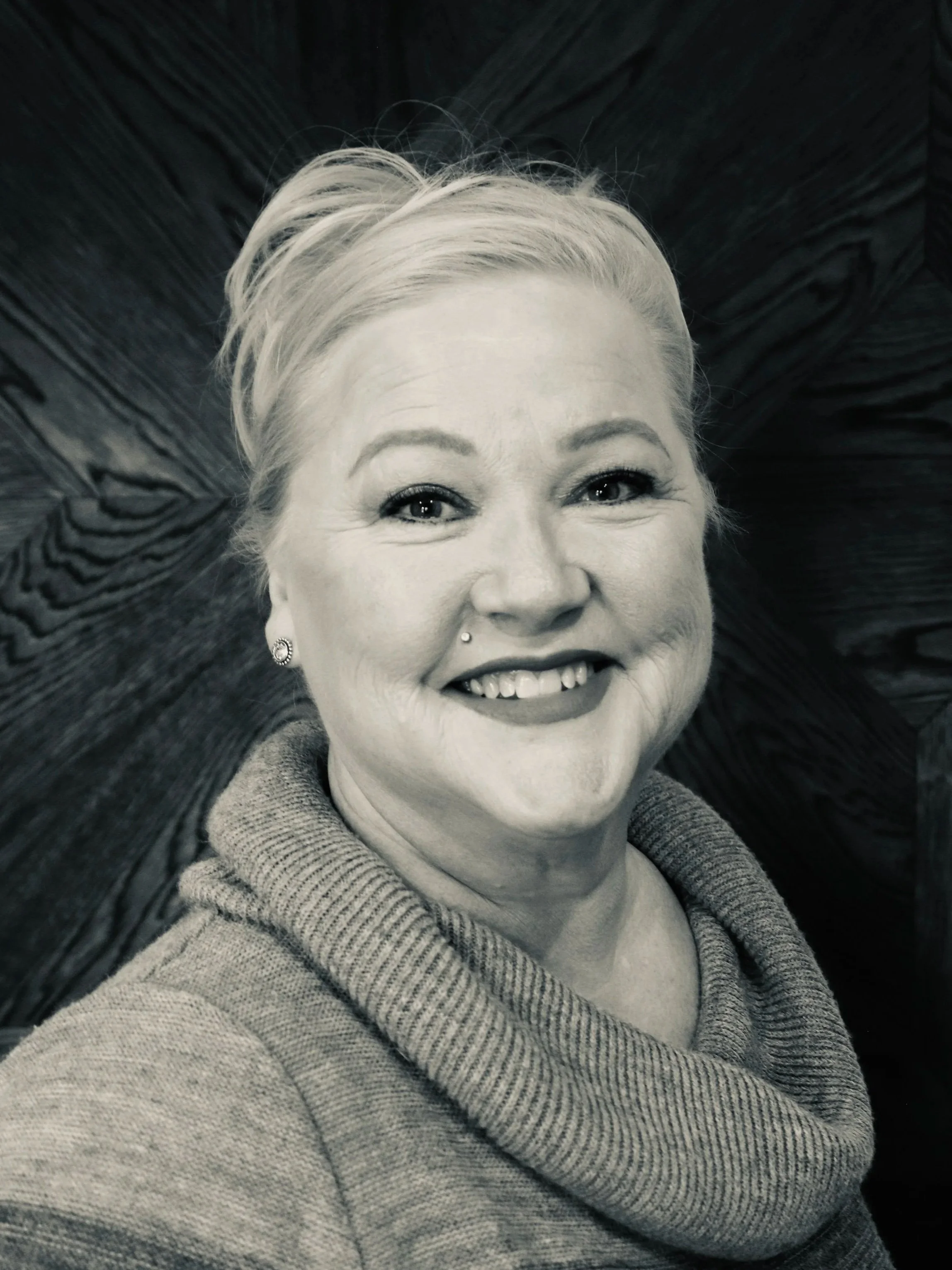 A woman with blonde hair smiling, wearing a gray hoodie, lying on a textured dark surface.
