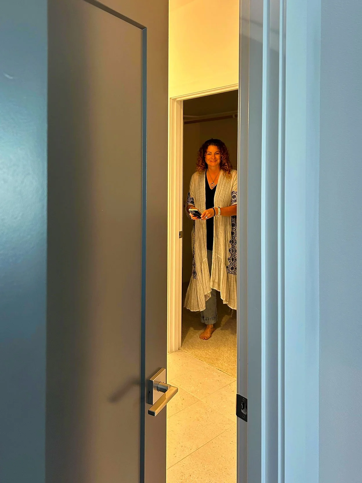 Woman standing in a doorway, smiling, holding a phone, wearing a long beige cardigan with embroidery, black top, and jeans, with barefoot on a beige carpet in Bend, Oregon.