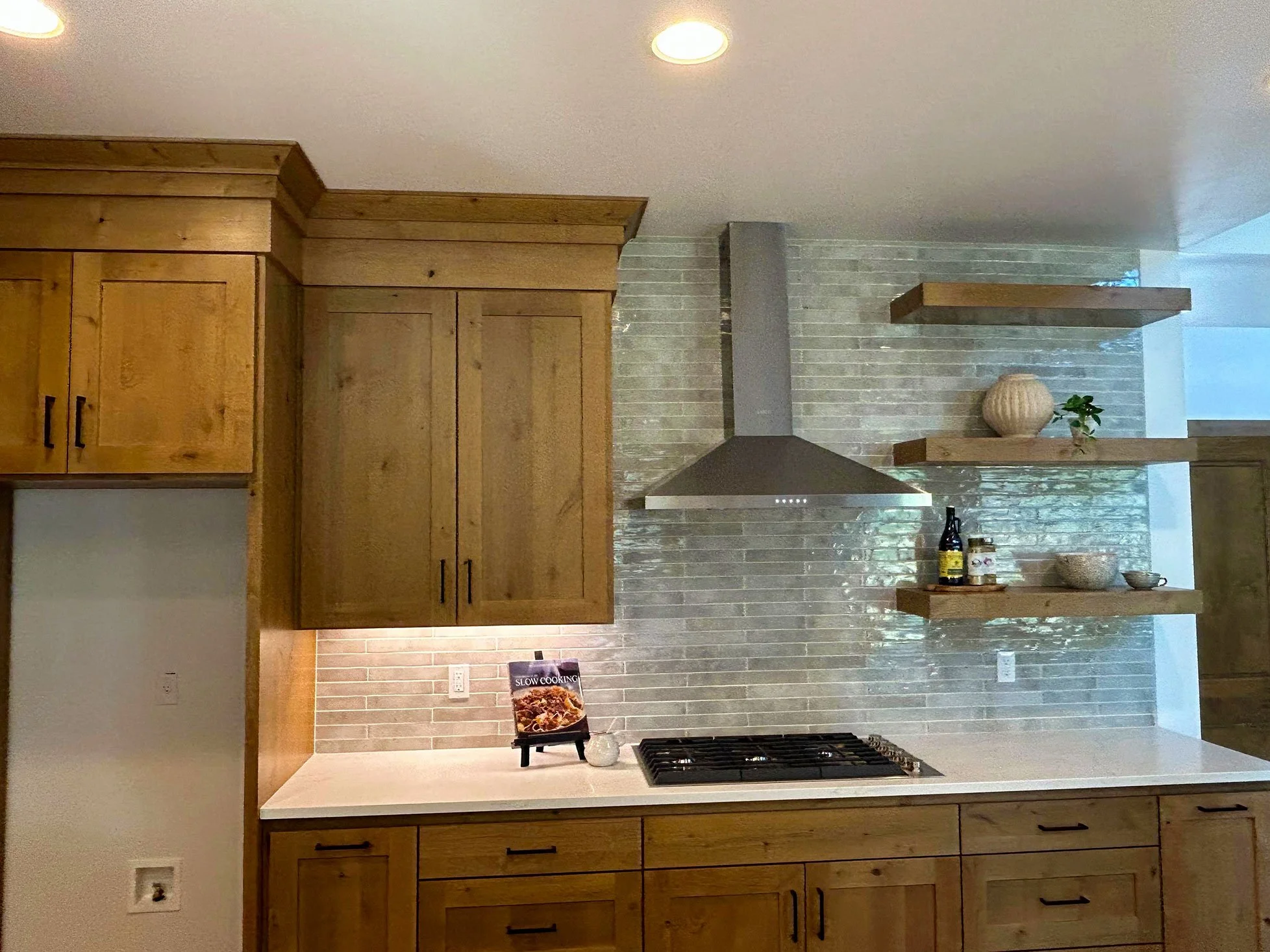 Kitchen with wooden cabinets, a white countertop, a stove, and open wooden shelves with decorative items on a gray brick backsplash in Bend, Oregon.