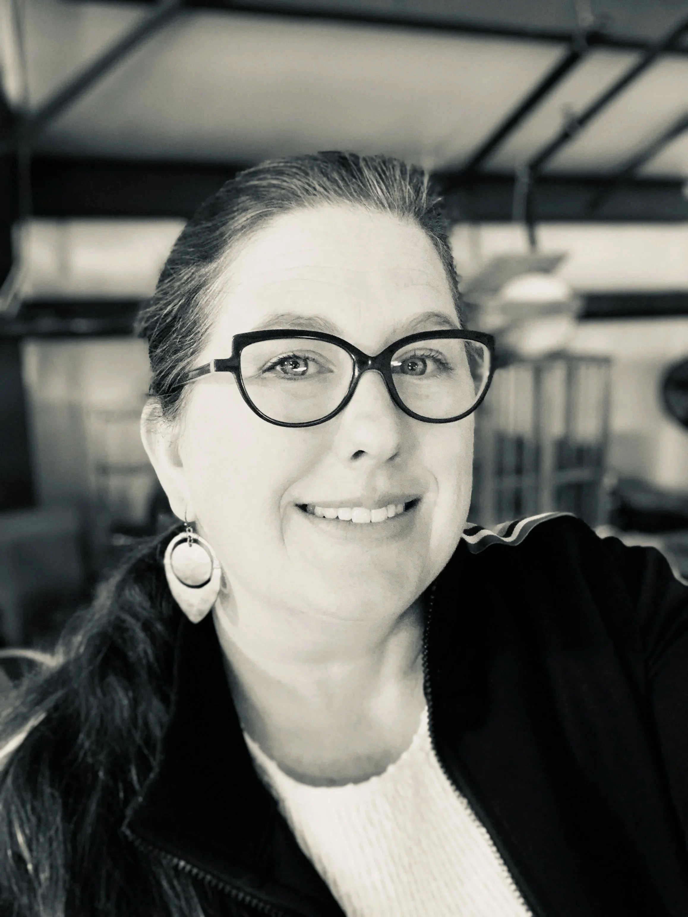 Close-up black and white photo of a smiling woman with glasses and earrings, wearing a black coat and white sweater, inside a building with metal shelving in the background.