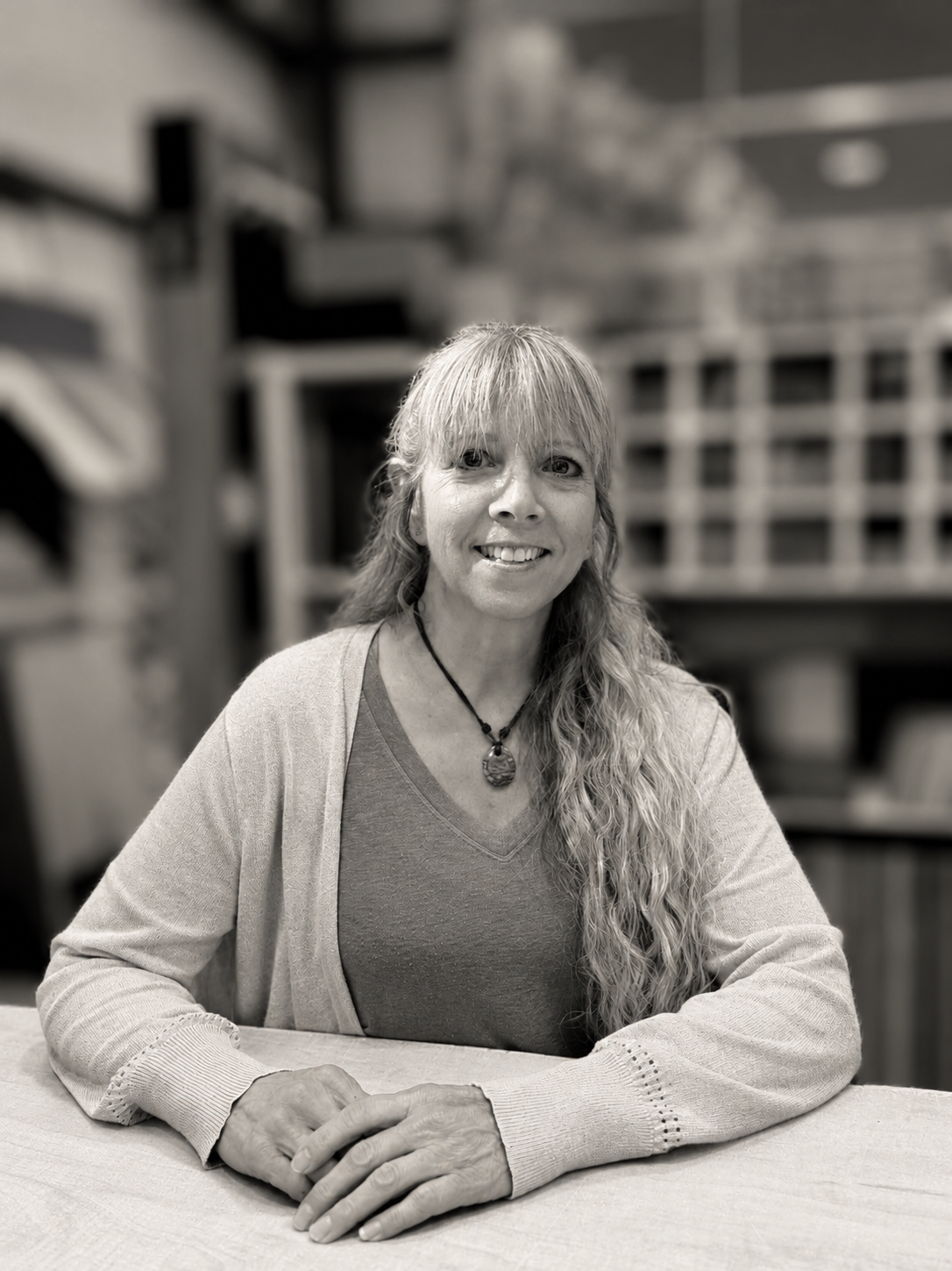 A woman with long wavy hair wearing a necklace and a light-colored cardigan sitting at a table in a workshop or office with shelves and equipment in the background.