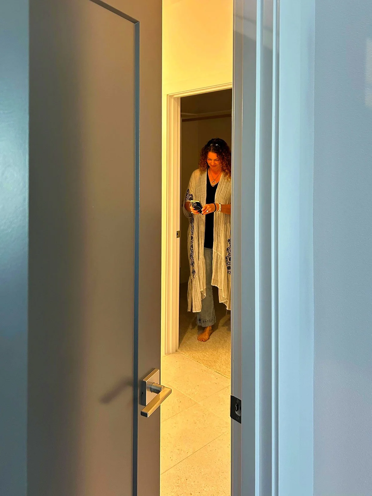 A woman with curly hair wearing a long cream-colored cardigan with blue embroidery, black top, and jeans, standing barefoot in a doorway looking at her phone in Bend, Oregon.