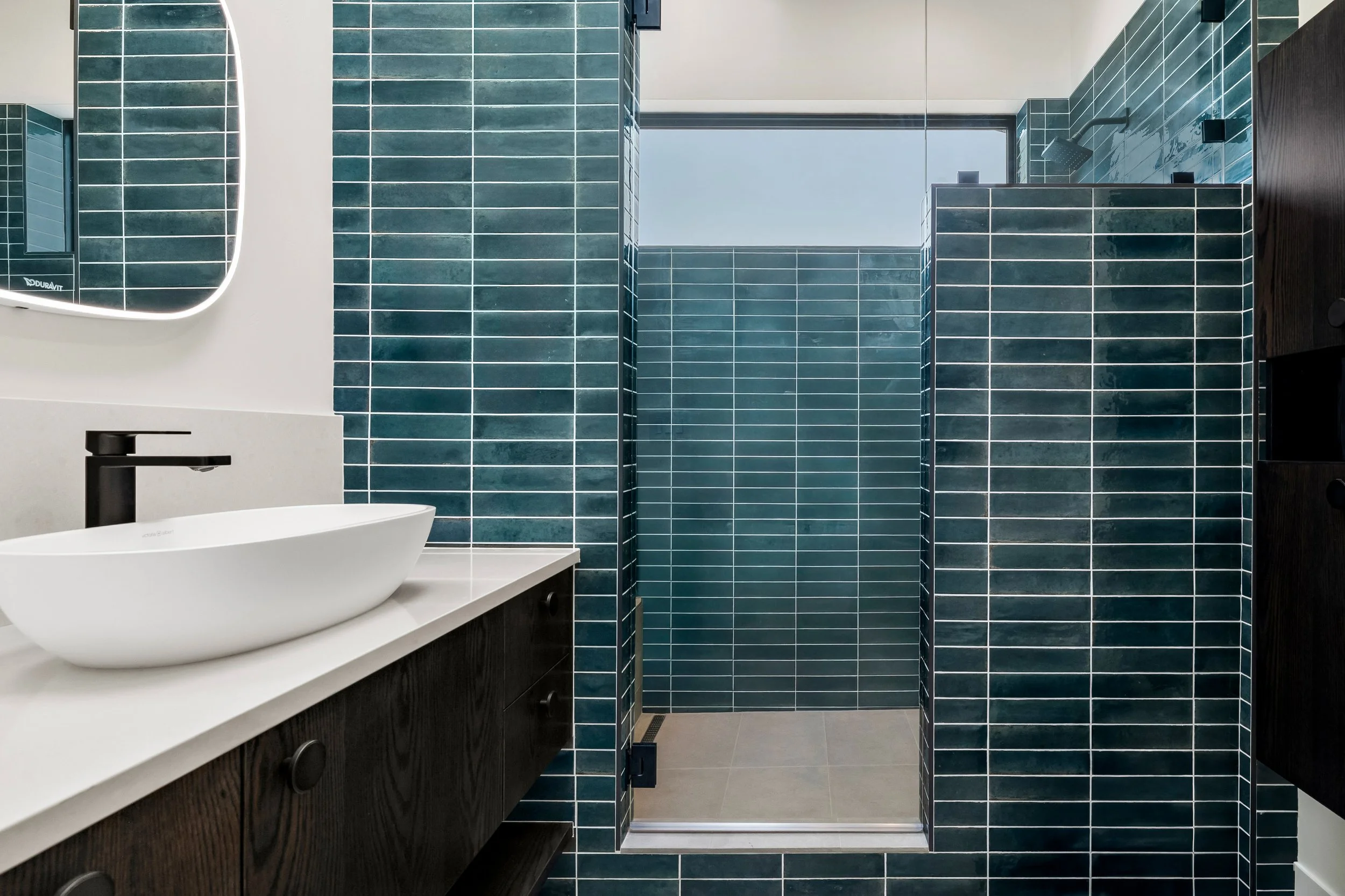Modern bathroom interior design with dark teal subway tiles, a white countertop with a vessel sink, and a separate shower area with a glass door.