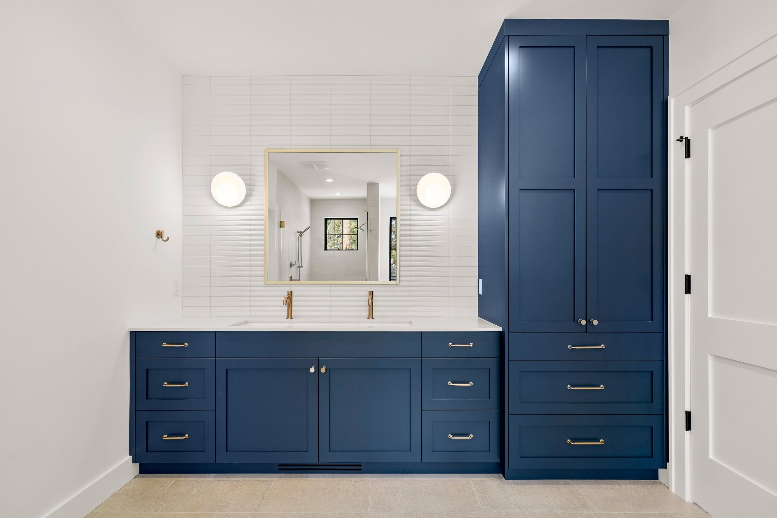 A modern bathroom vanity with navy blue cabinets, gold hardware, a white countertop, and a large mirror with two wall-mounted lights. The bathroom interior design has white walls and a tiled floor.