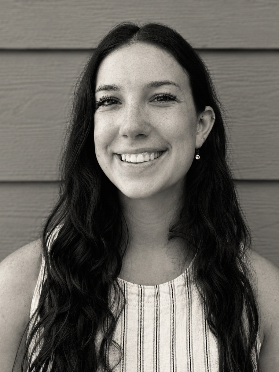 Black and white portrait of a young woman with long wavy dark hair, smiling, wearing earrings and a sleeveless striped top, standing in front of a wooden wall.