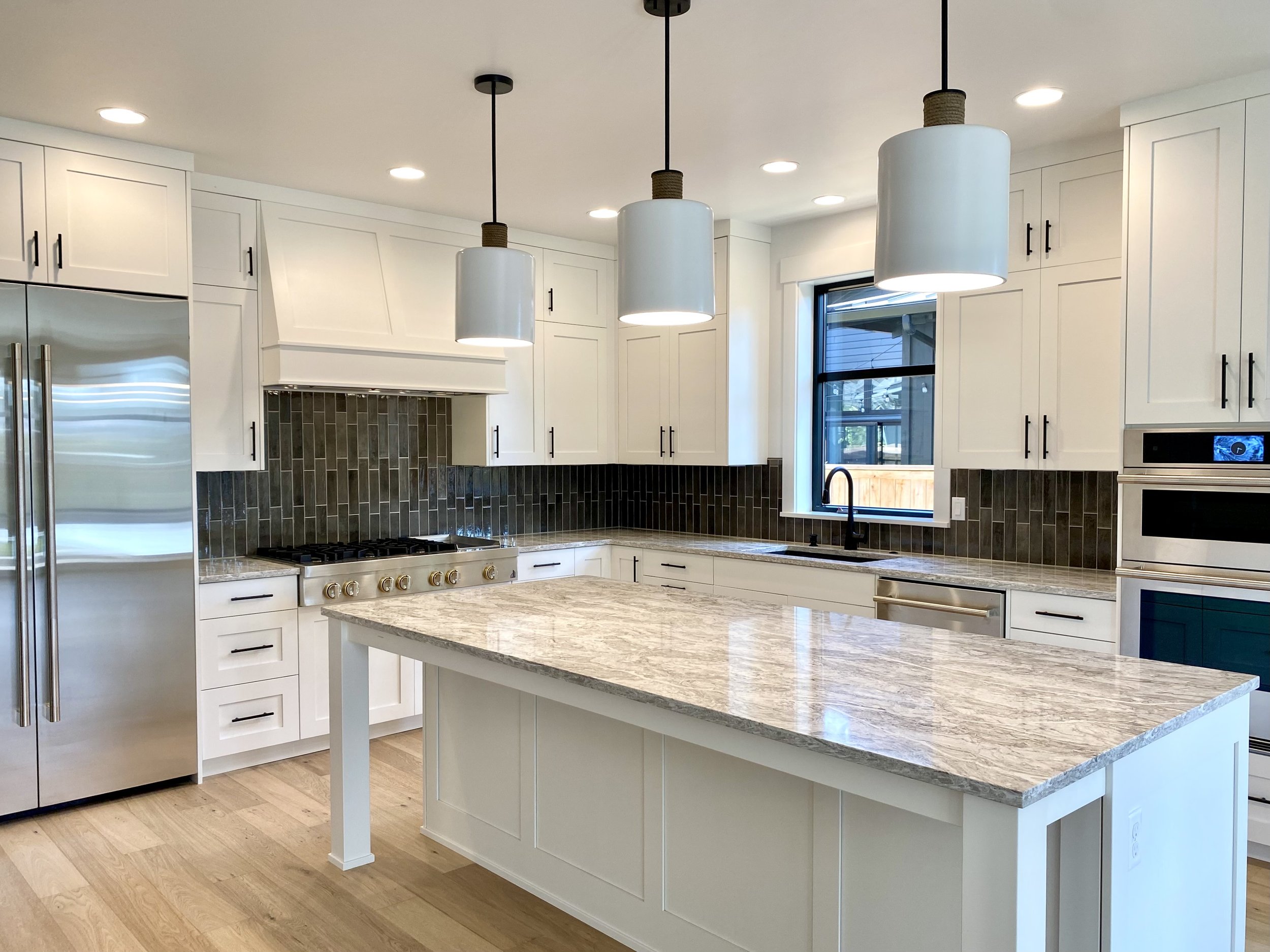 Modern kitchen with white cabinetry, a central island with a granite countertop, stainless steel refrigerator, black tile backsplash, black hardware, and pendant lights in Bend, Oregon.