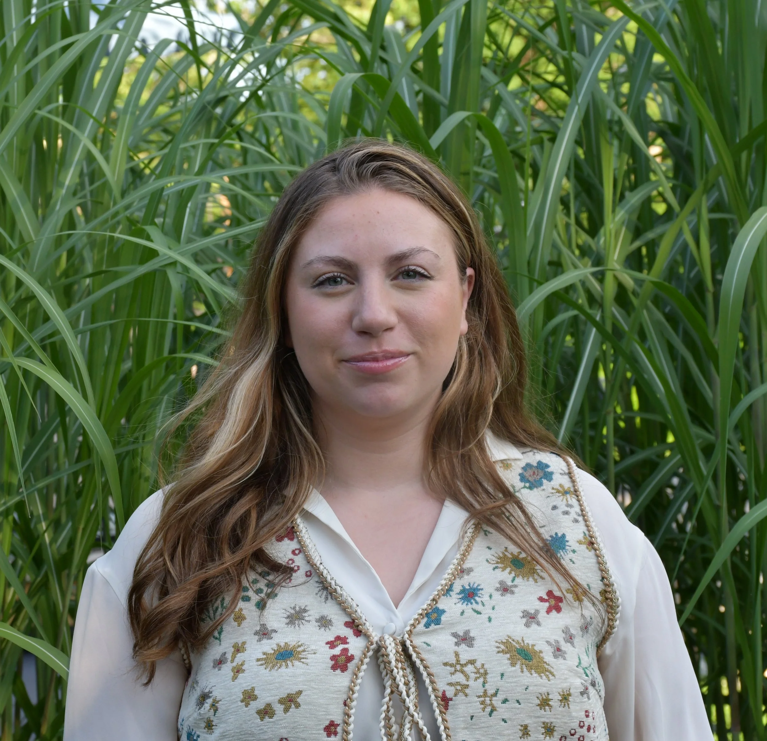 Portrait of a young woman with long curly hair standing outdoors in front of tall green grass or plants, wearing a decorative embroidered white blouse with floral patterns and a pearl necklace.