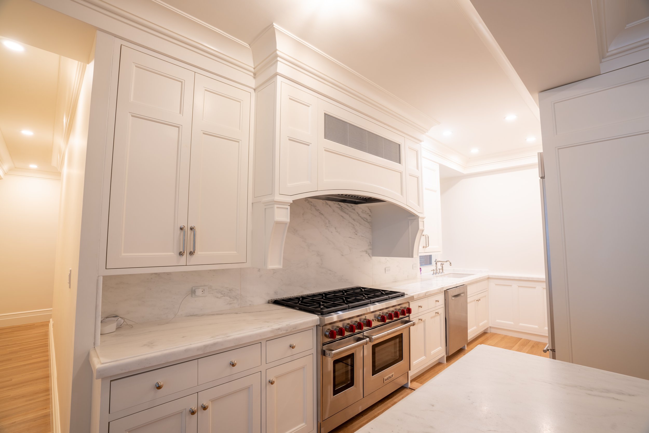 Modern kitchen with white cabinets, marble countertops, stainless steel appliances, including a range and dishwasher, and recessed lighting.