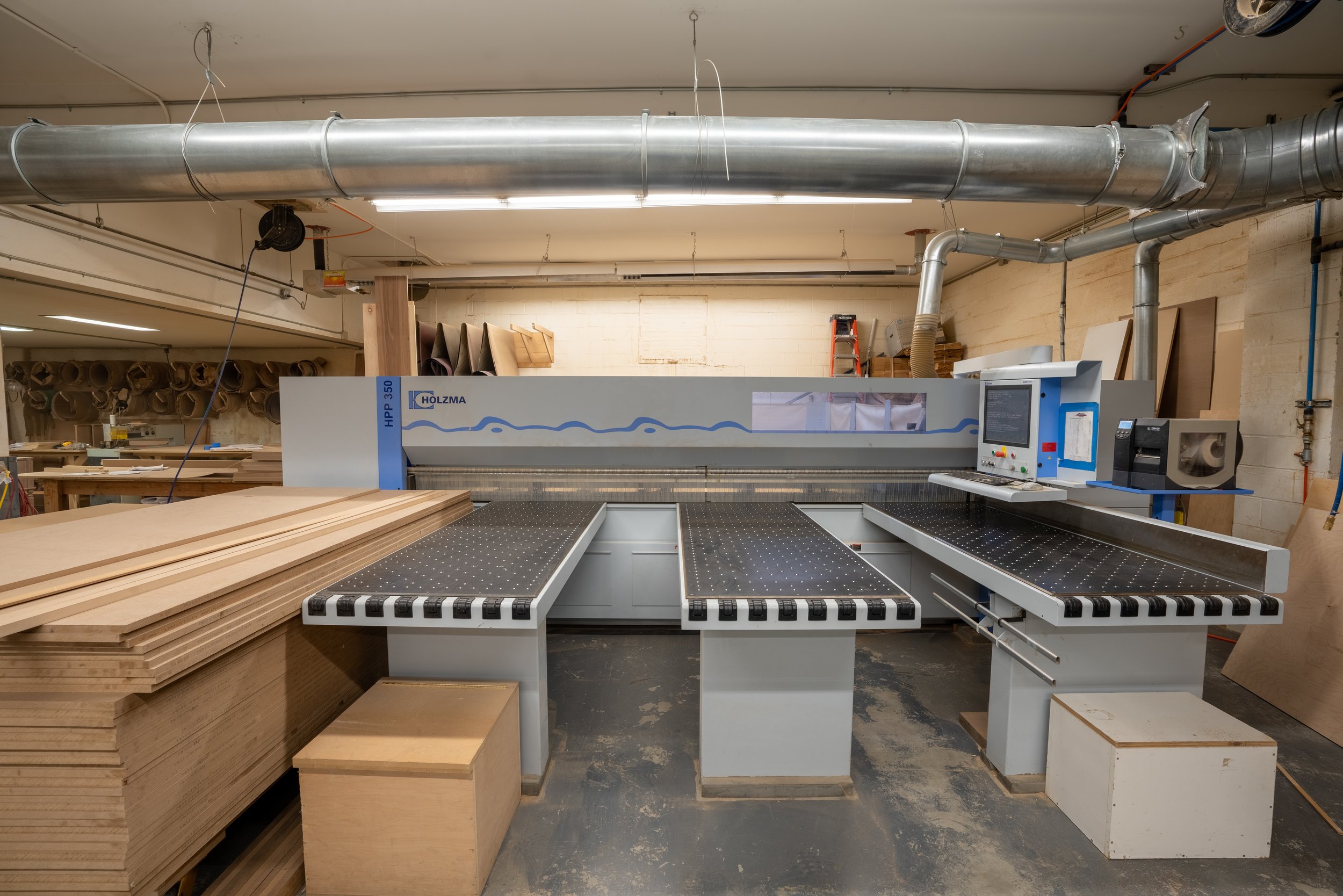 Woodworking factory with industrial cutting machine, Holzma HPP 350, stacks of plywood, ventilation ducts, and control panel in a workshop setting.