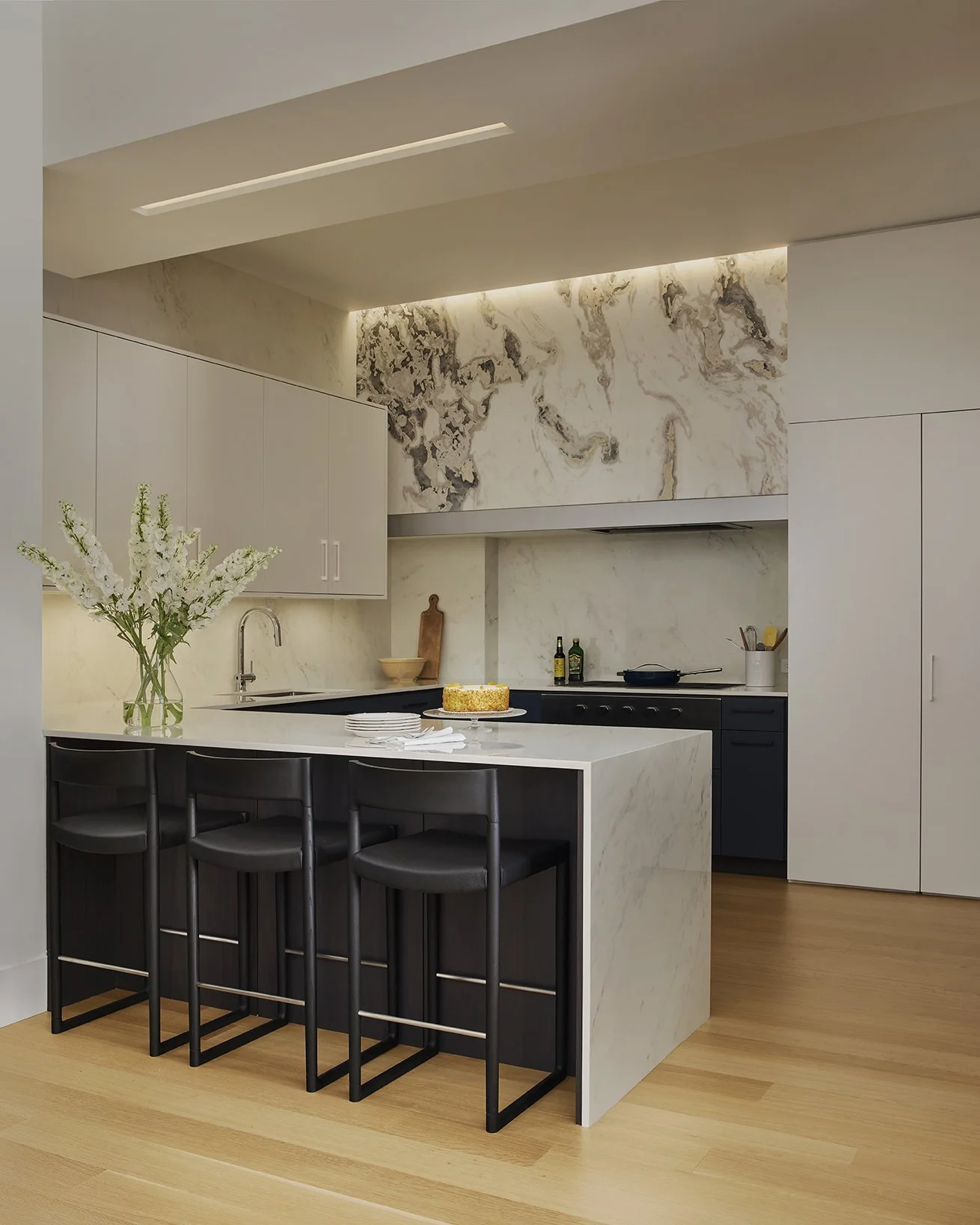 Modern kitchen with white marble countertops and backsplash, black bar stools, a vase of white flowers, and a birthday cake on the island.