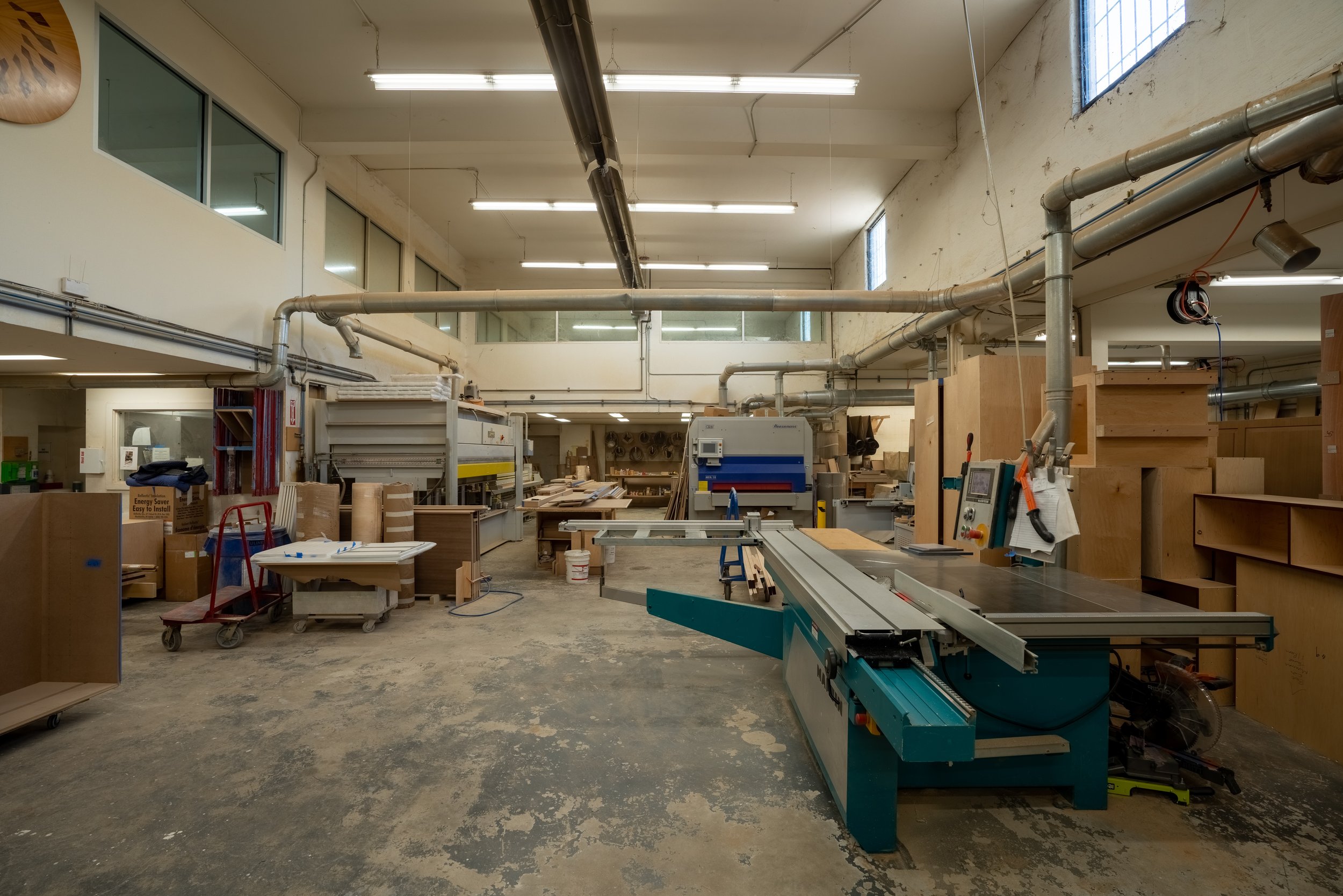 Interior of a woodworking workshop with various machines and tools, unfinished wooden furniture, and large ventilation pipes.