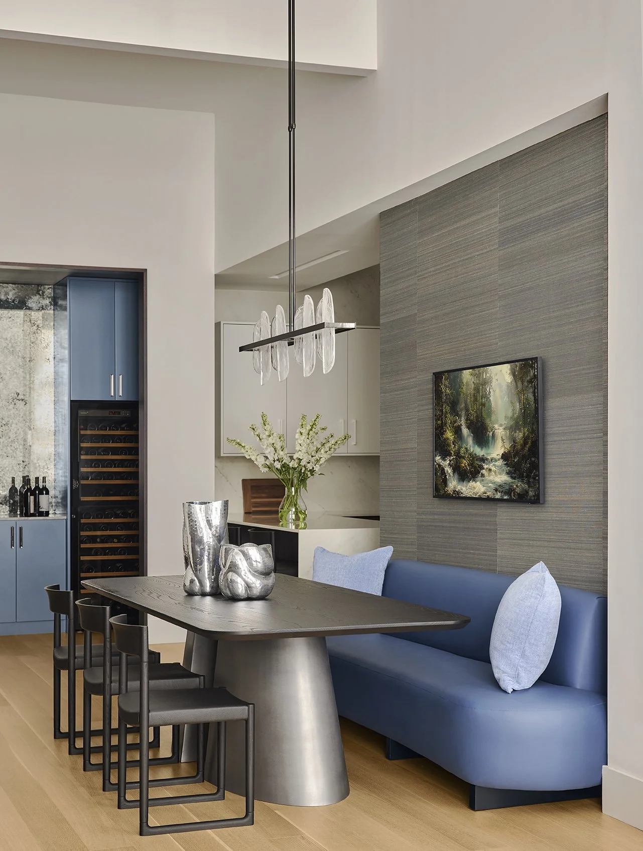 Modern dining area with black table, blue booth with white pillows, silver vases, and cream chairs. In the background, there is a kitchen with white cabinets, a vase with flowers, and a landscape painting on the wall.