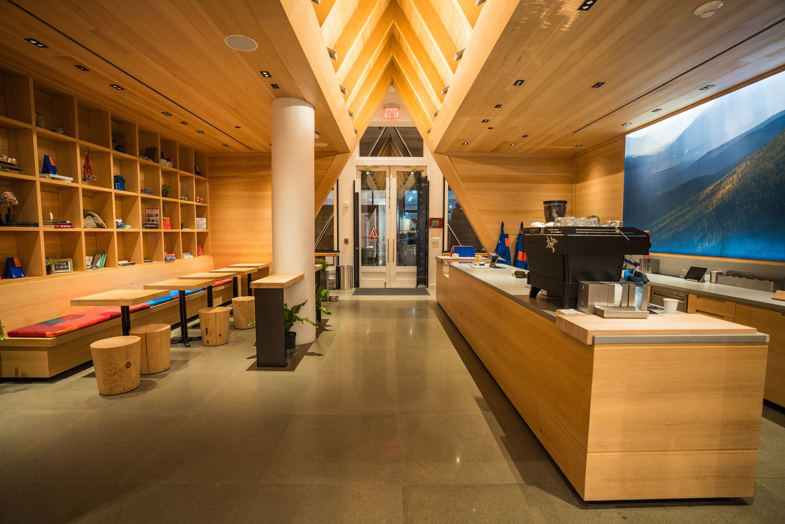 Modern coffee shop interior with wood accents, featuring seating with cushions, a long counter with a coffee machine, and shelves with books and decor items.