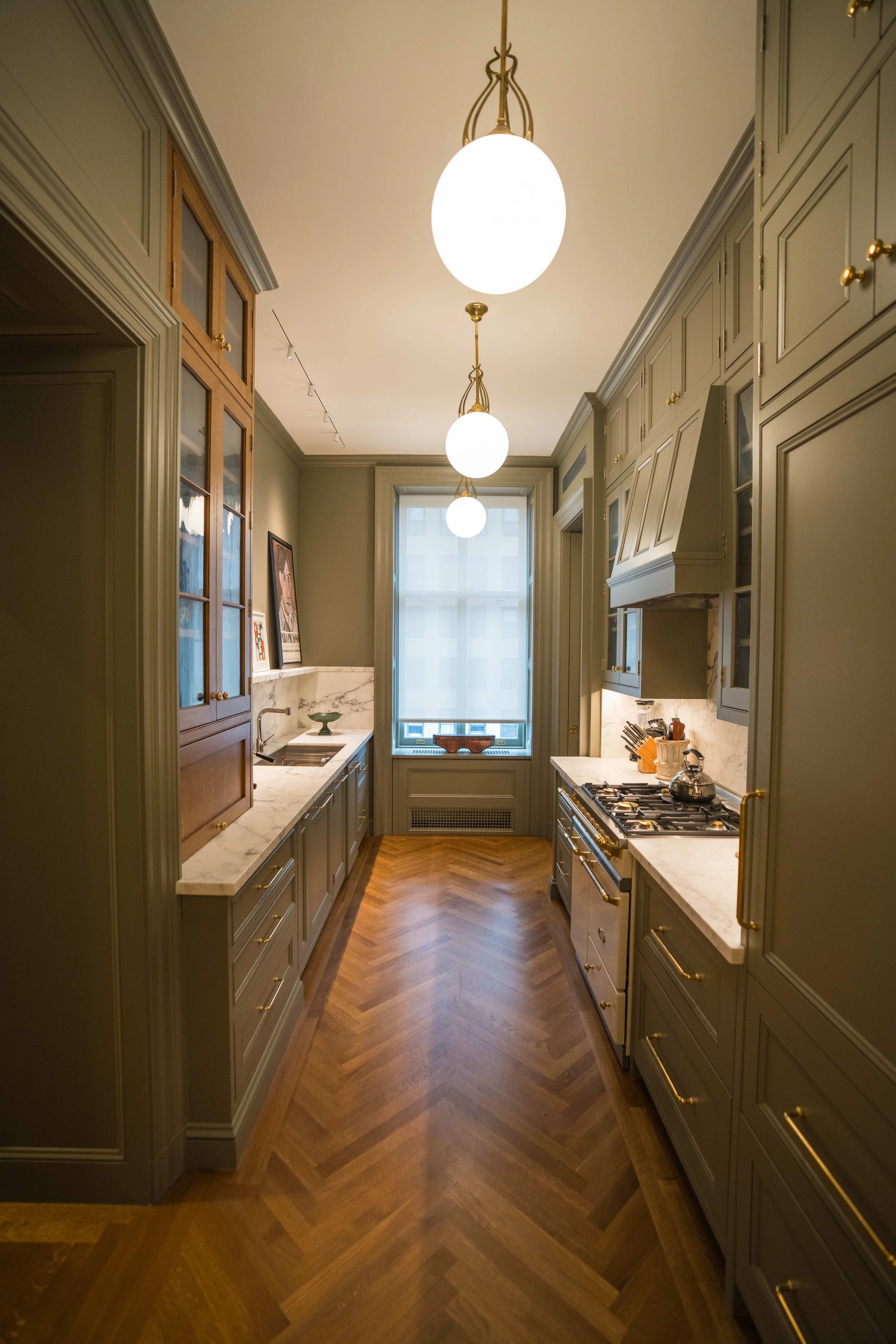 A narrow kitchen with sage green cabinets, gold hardware, marble countertops, a window with natural light, and hanging globe pendant lights.