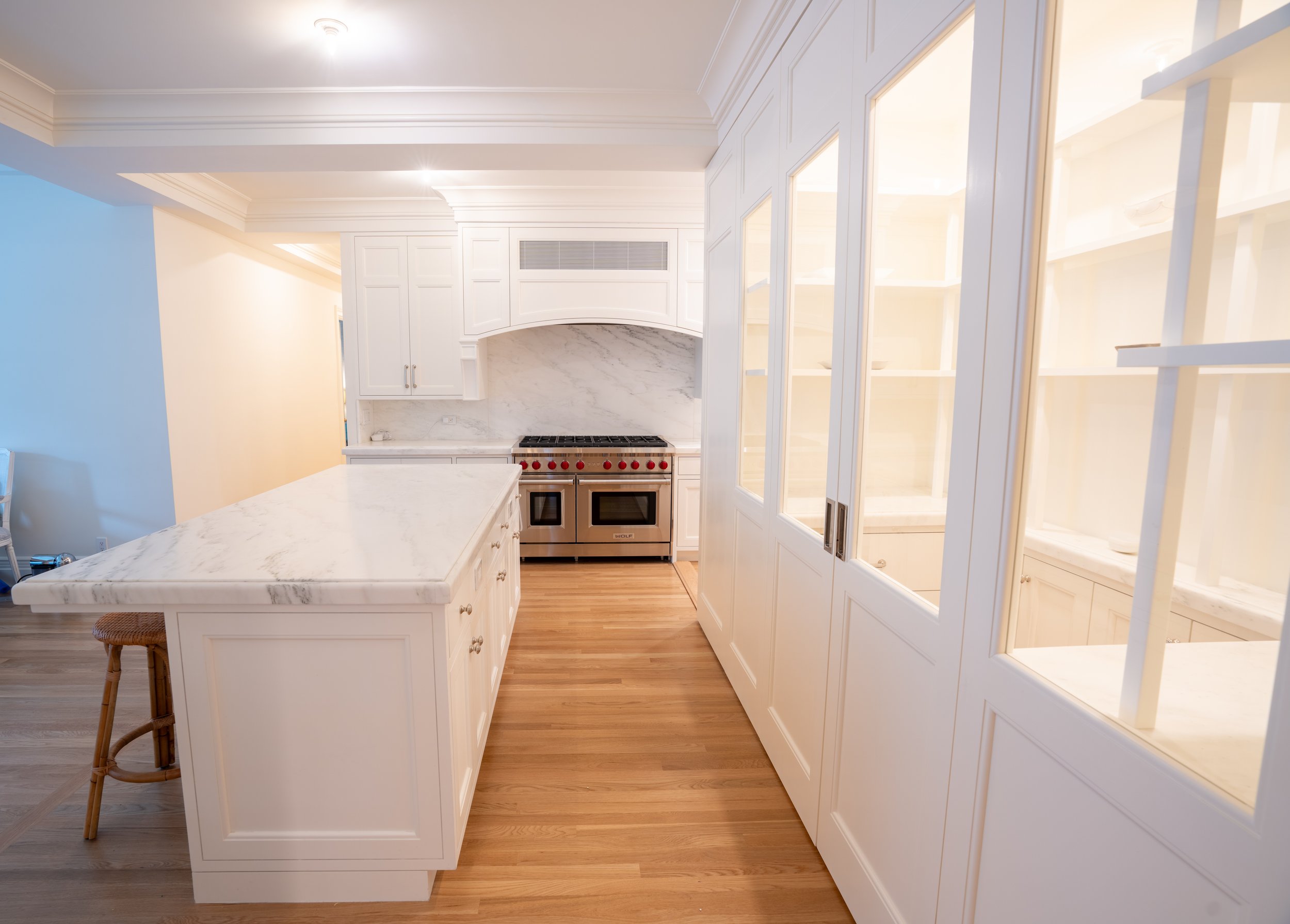 Modern kitchen with white cabinetry, marble countertops, stainless steel stove, wood flooring, and a glass-paneled pantry door.