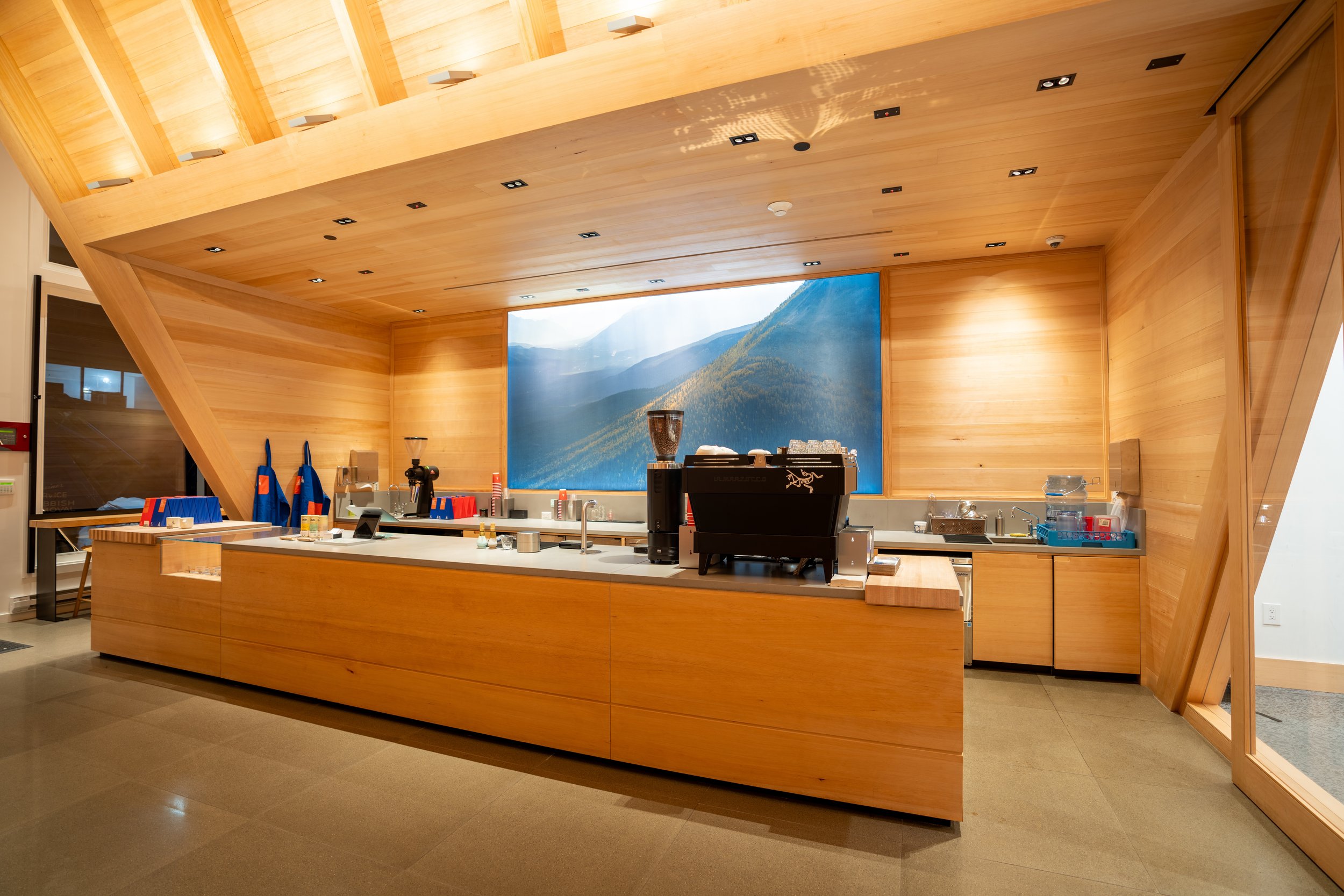 Modern coffee shop counter with wooden design, featuring a large coffee machine, grinders, and various utensils. A mural of a mountain landscape is displayed on the wall behind.