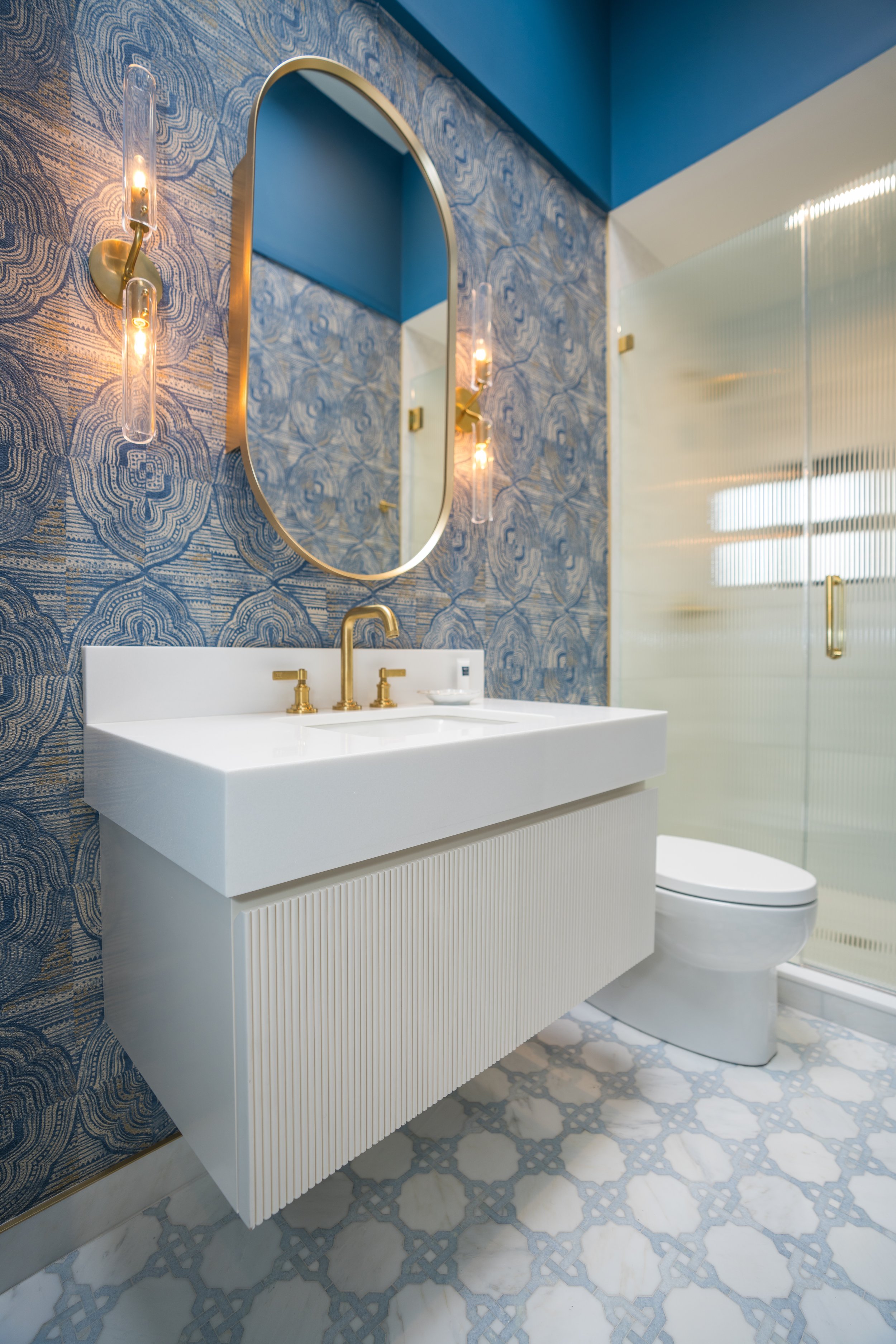 Modern bathroom with blue textured wallpaper, white floating vanity with gold faucets, oval mirror, decorative wall lights, and patterned tile flooring.
