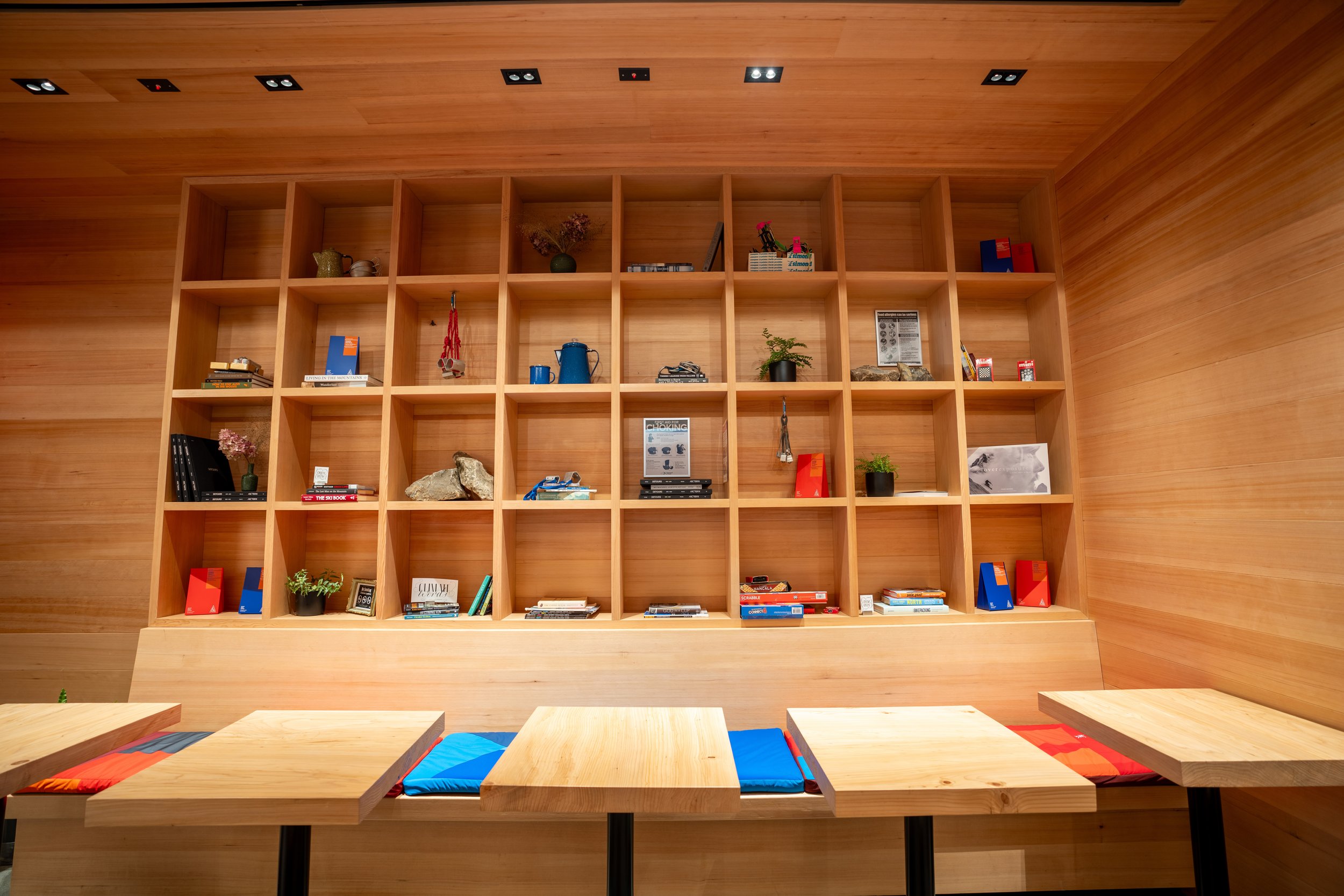 Wooden shelving with various items, books, and decor in a modern interior space, with wooden tables and colorful cushions in the foreground.
