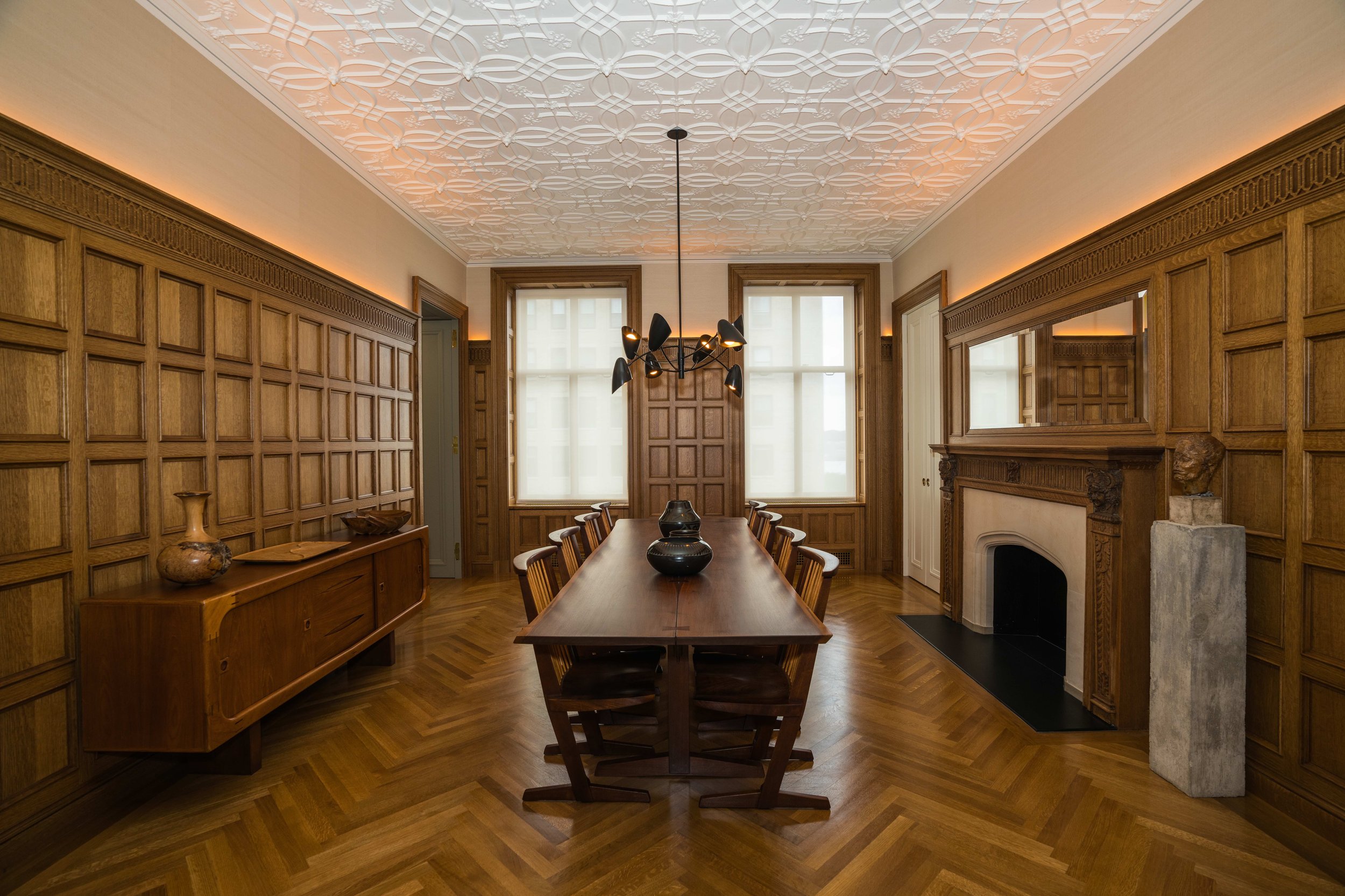A wood-paneled dining room with a long table and chairs, a fireplace with an ornate mantel, a modern chandelier, and large windows.