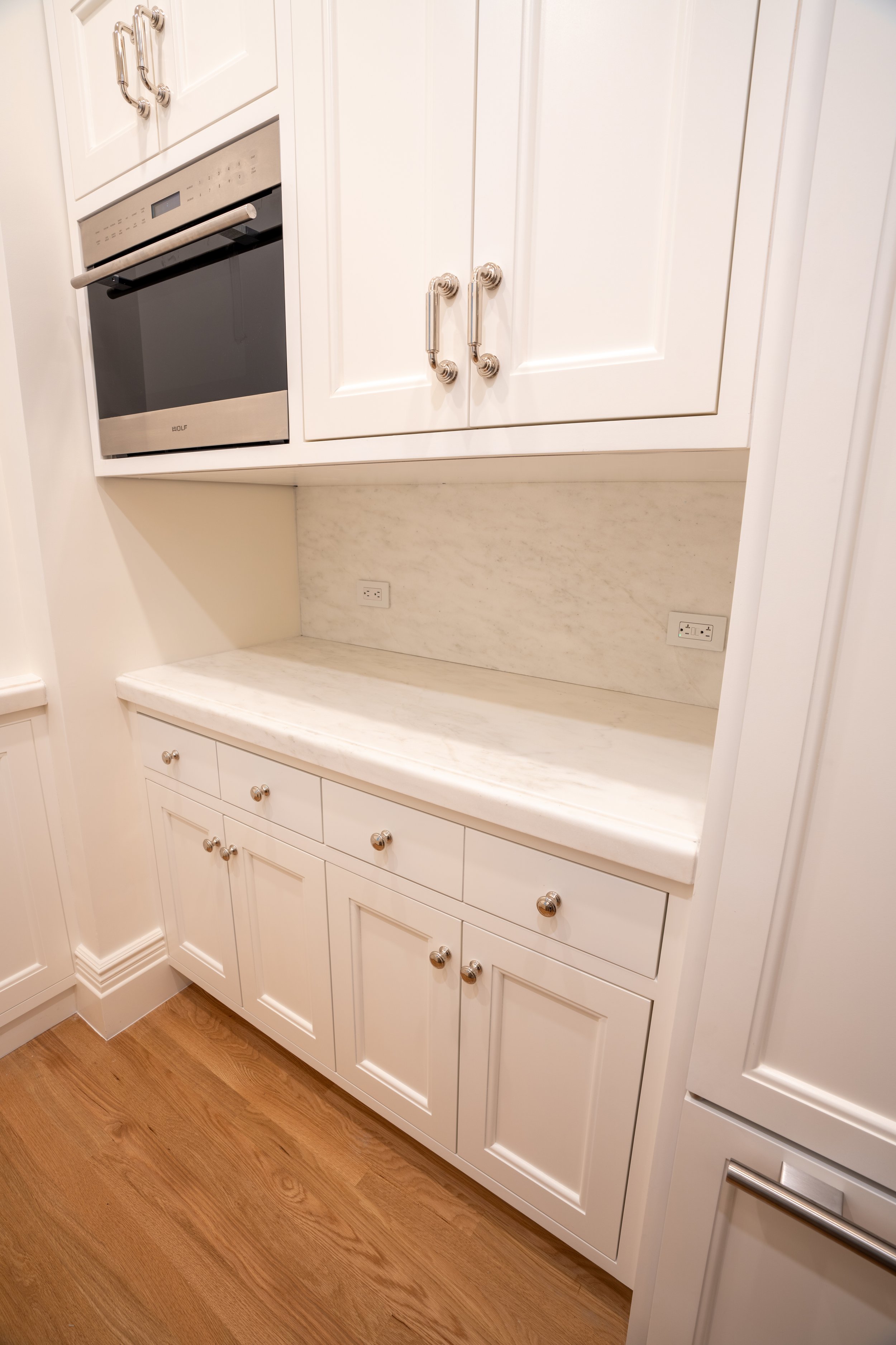 White kitchen cabinets and drawers with a built-in stainless steel oven, marble countertops, and wood flooring.