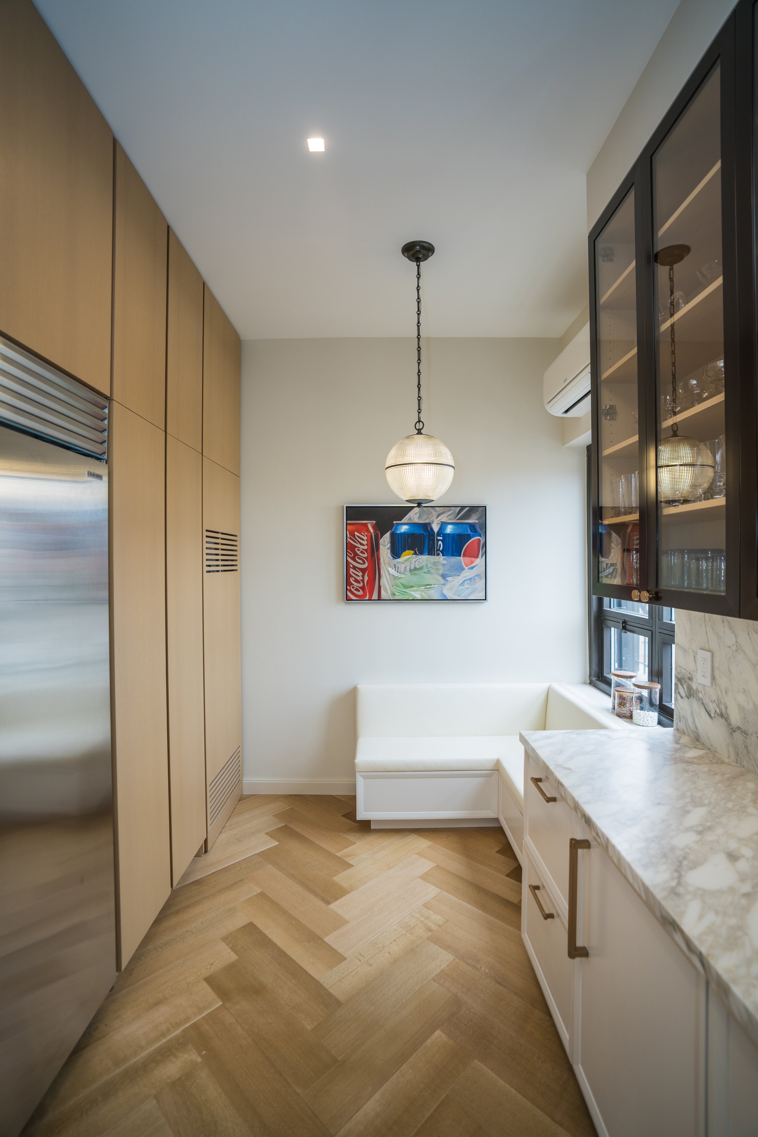 Modern kitchen with herringbone wooden floor, light wood cabinets, white marble countertop, and a corner bench. A spherical pendant light hangs from the ceiling. A framed artwork featuring beverage cans is on the wall.