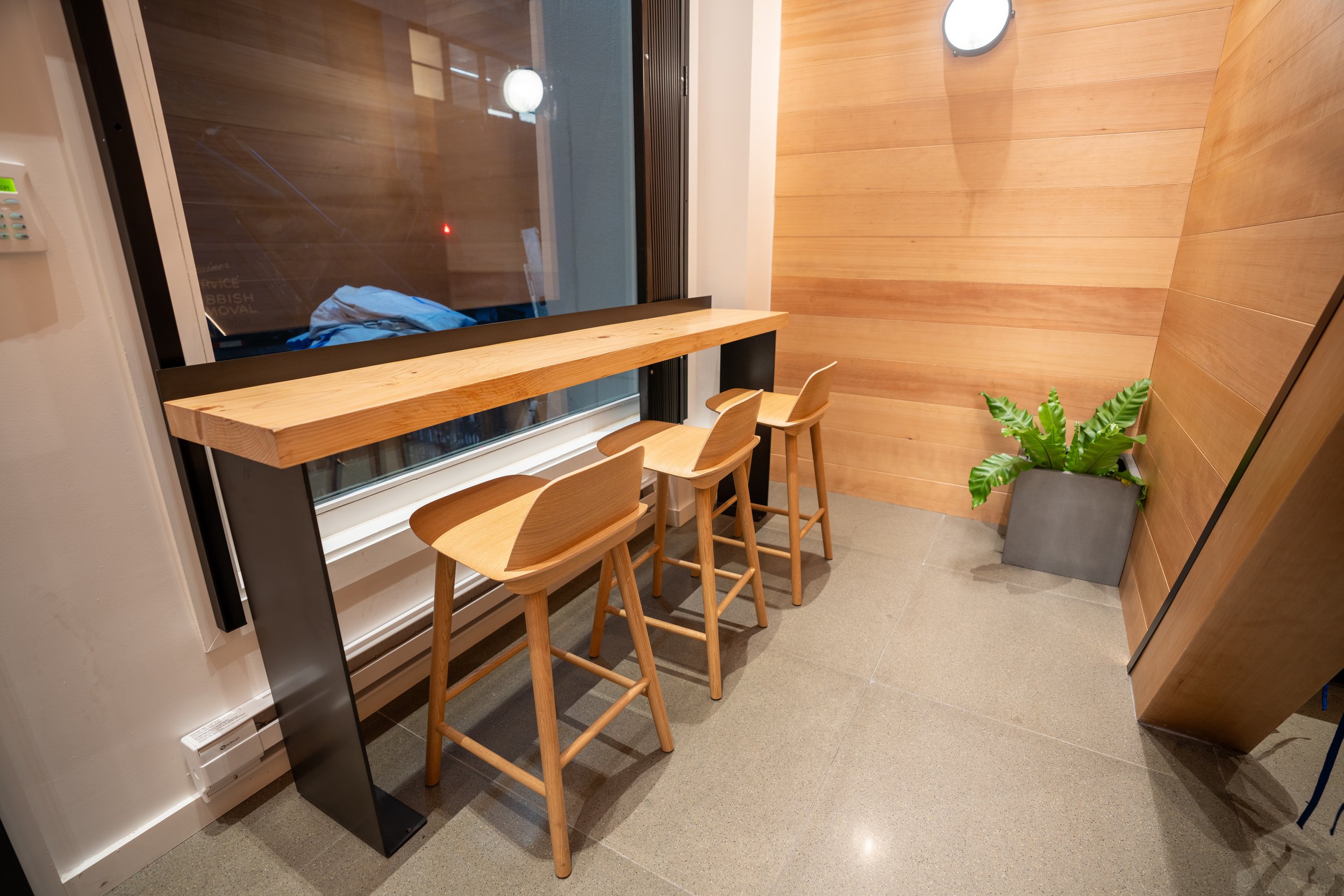 Modern seating area with a tall wooden table and three stools next to a window. The area has a polished tile floor, wooden wall panels, and a potted plant.