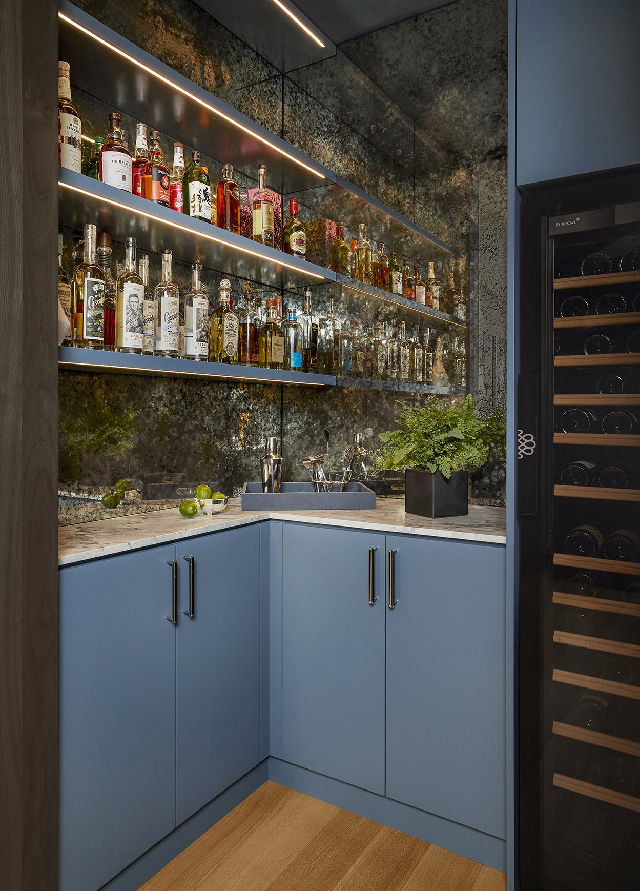 A modern bar area with a marble countertop, blue cabinetry, a gray sink, and a backsplash with bottles of liquor on illuminated glass shelves. There is a plant in a black pot and limes on the counter, with a wine cooler to the right.