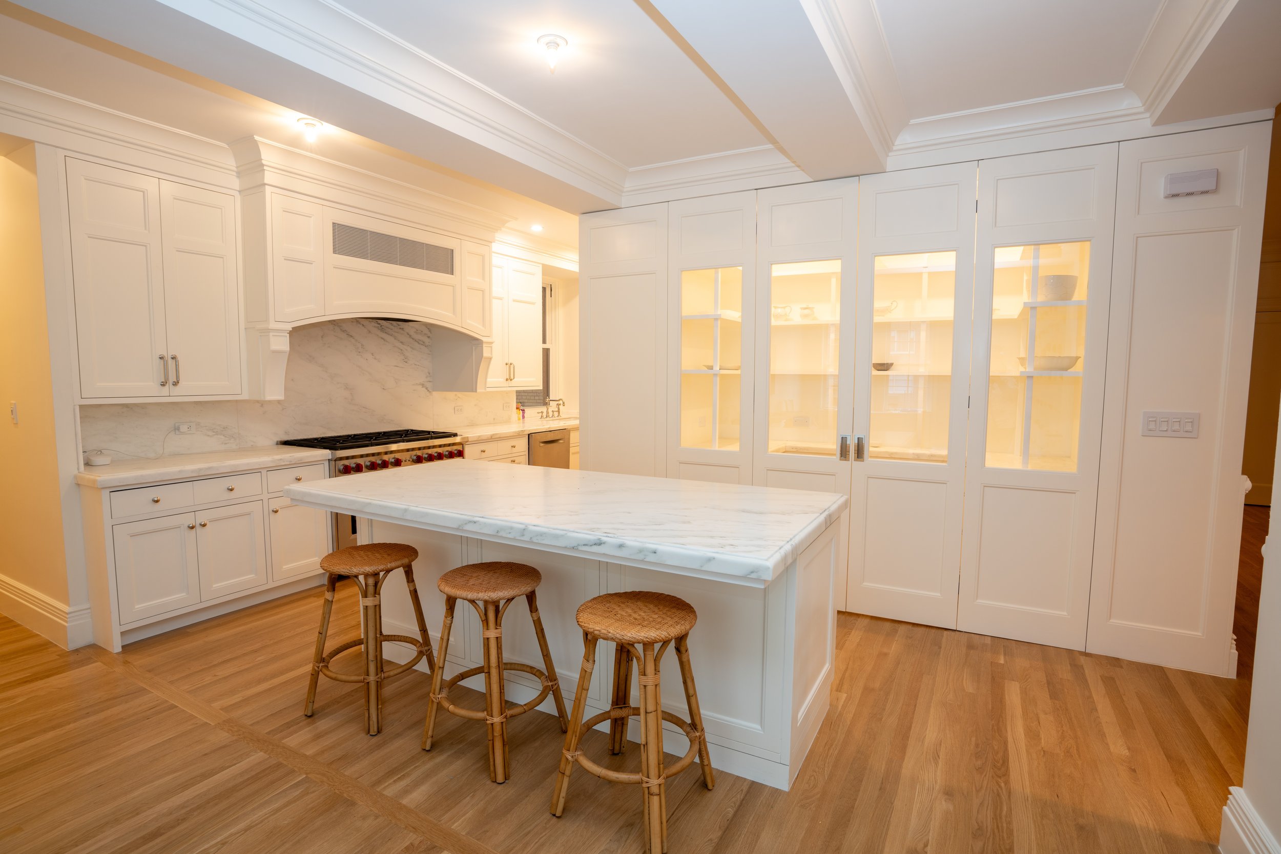 Modern kitchen with white cabinetry, marble countertops, and wooden floor. Features an island with wicker stools, built-in appliances, and glass-front cabinets.