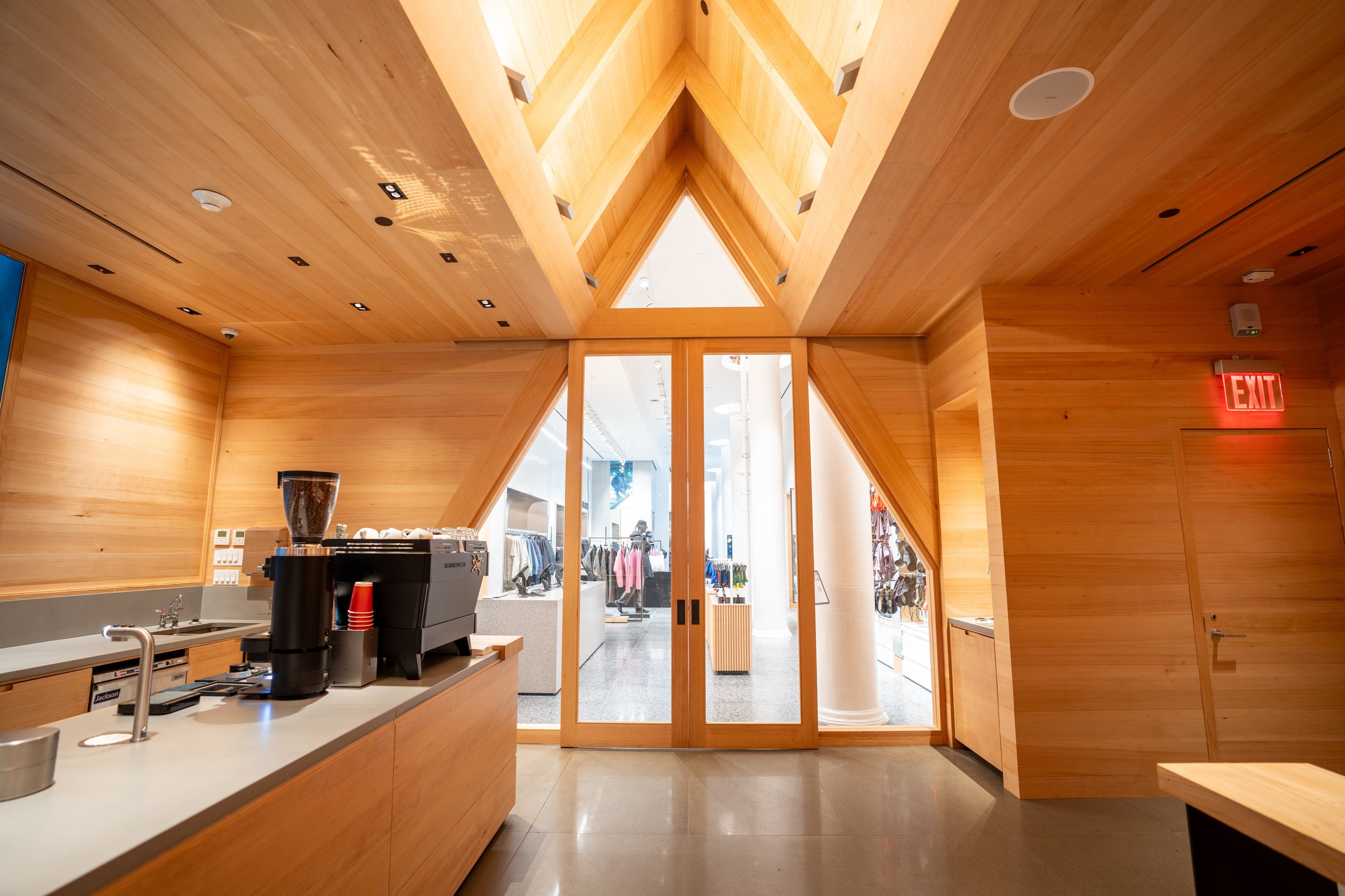 Interior of a modern coffee shop with wooden walls and a ceiling leading to a glass door entrance. A coffee machine sits on the counter, and clothing is visible through the doors, suggesting a retail store combination.