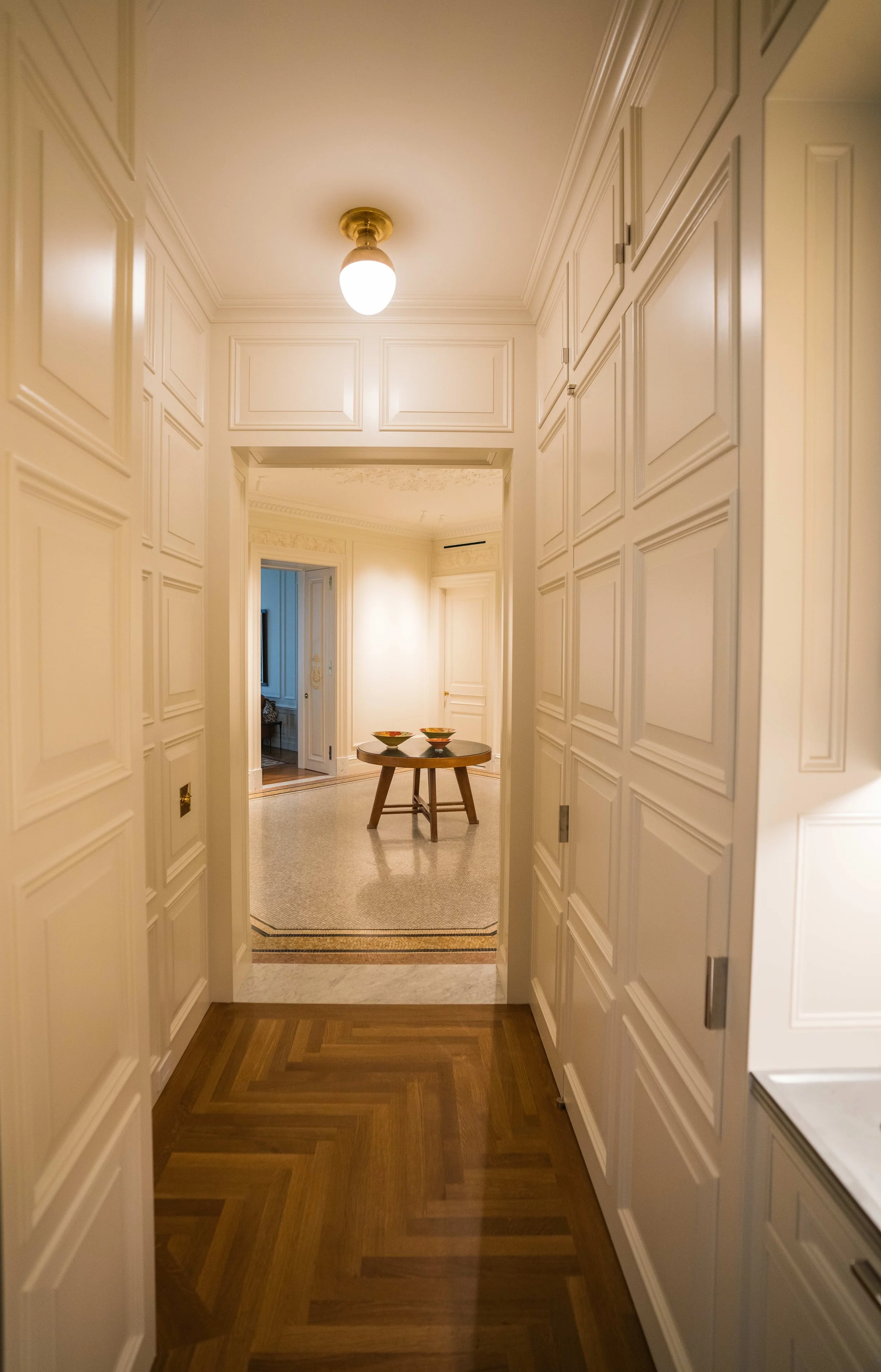 A hallway with white paneled walls, a ceiling light, and a wooden floor. At the end of the hallway is a small round table with decorative bowls, leading into another room with visible furniture.