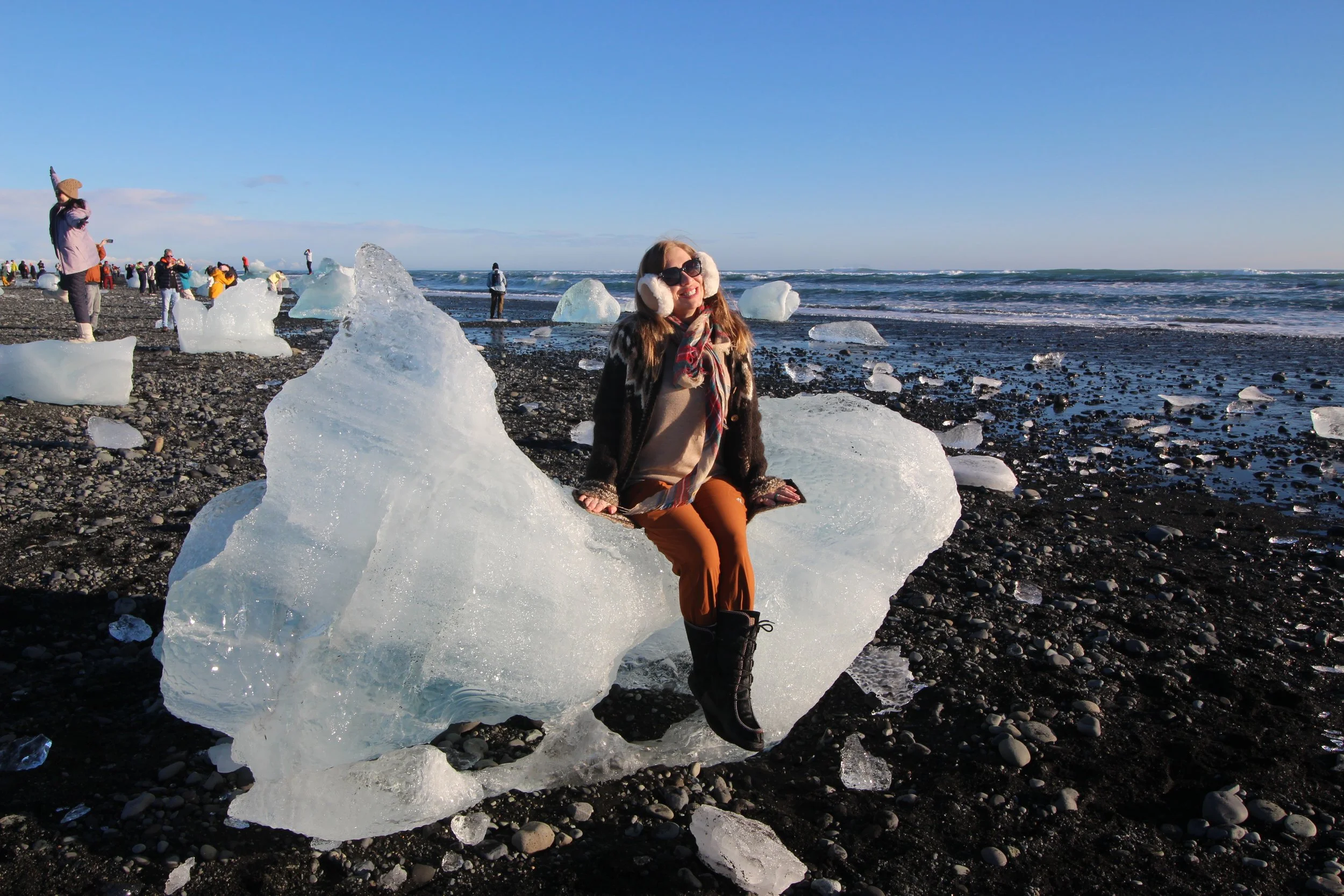 A woman sitting on a large chunk of ice on a rocky beach, smiling and wearing sunglasses, a scarf, earmuffs, and boots. In the background, people are gathered along the shoreline, with some standing on smaller ice formations, and the ocean waves are visible under a clear blue sky.