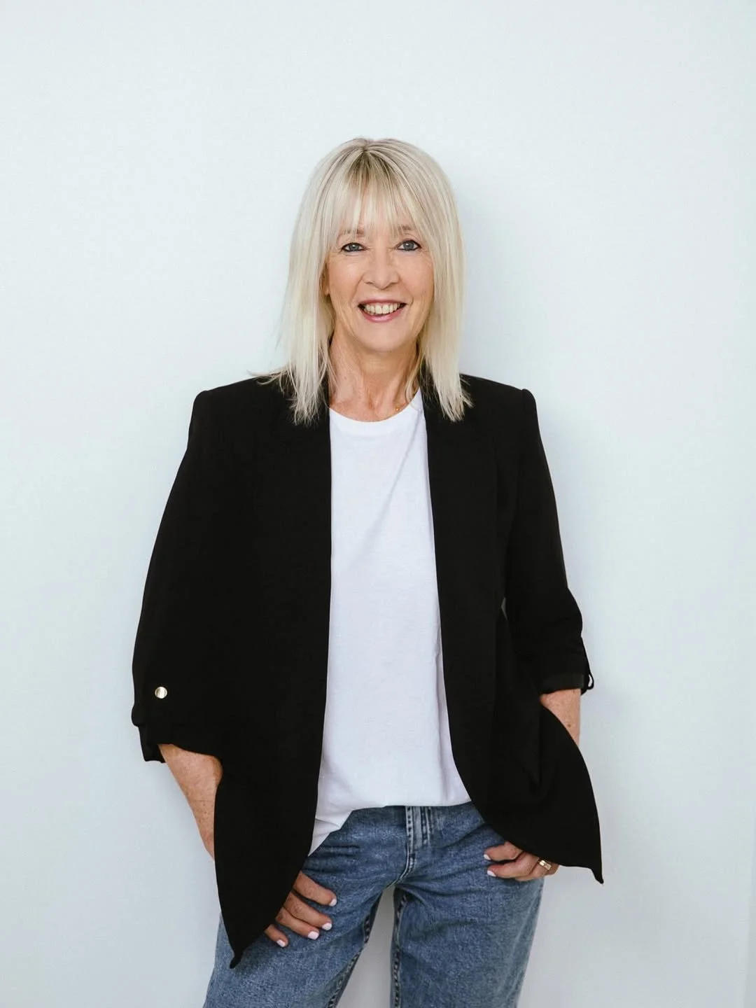 A middle-aged woman with blonde hair, wearing a black blazer, white t-shirt, and blue jeans, standing against a plain white background and smiling.