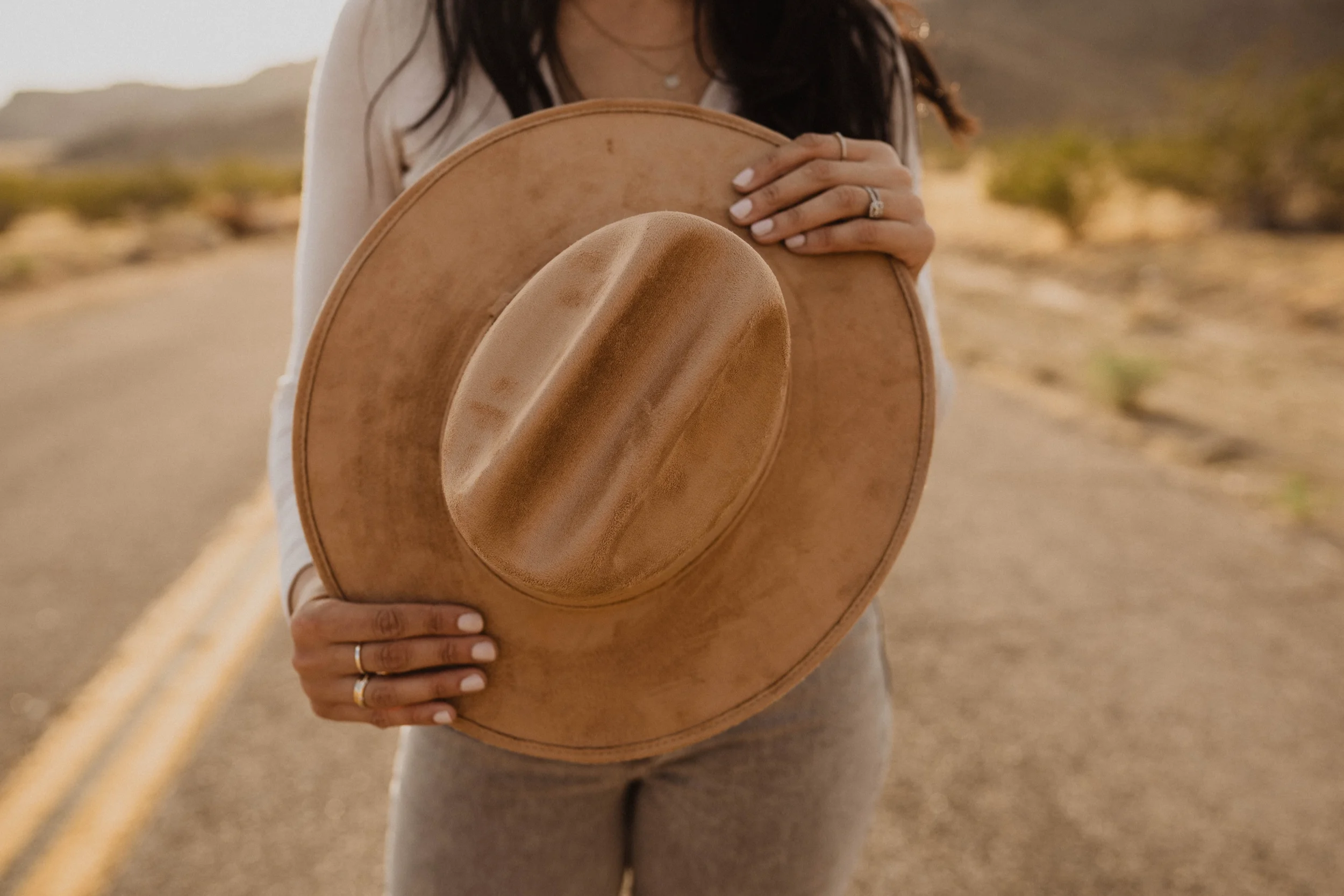 A woman standing on a desert road holding a tan cowboy hat in front of her face. She has long dark hair, and is wearing rings on her fingers, a beige top, and beige pants. The background shows an arid landscape with mountains and sparse vegetation.
