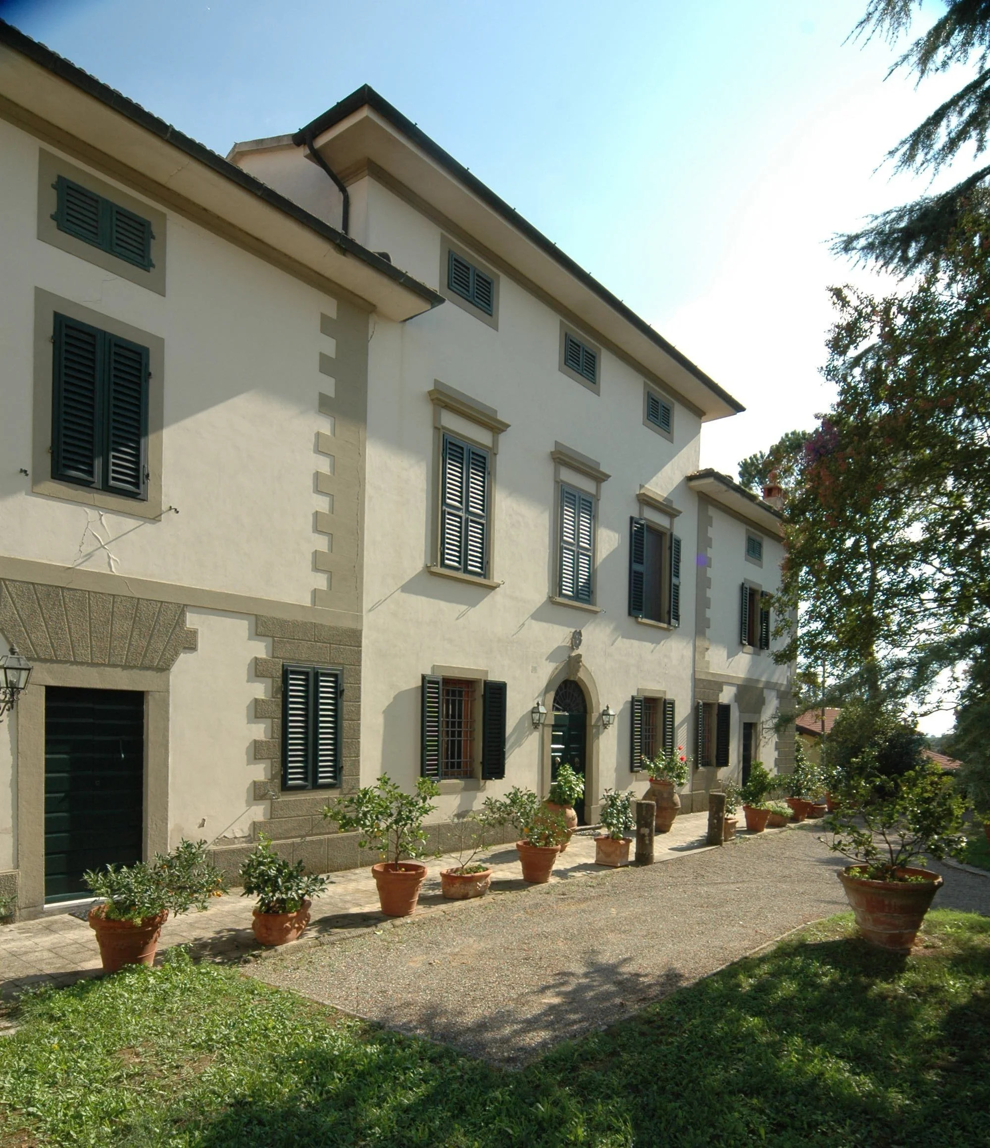 Casa bianca con finestre con persiane nere e piante in vaso lungo la parete di fronte all'ingresso, strada di ghiaia e alberi sullo sfondo.