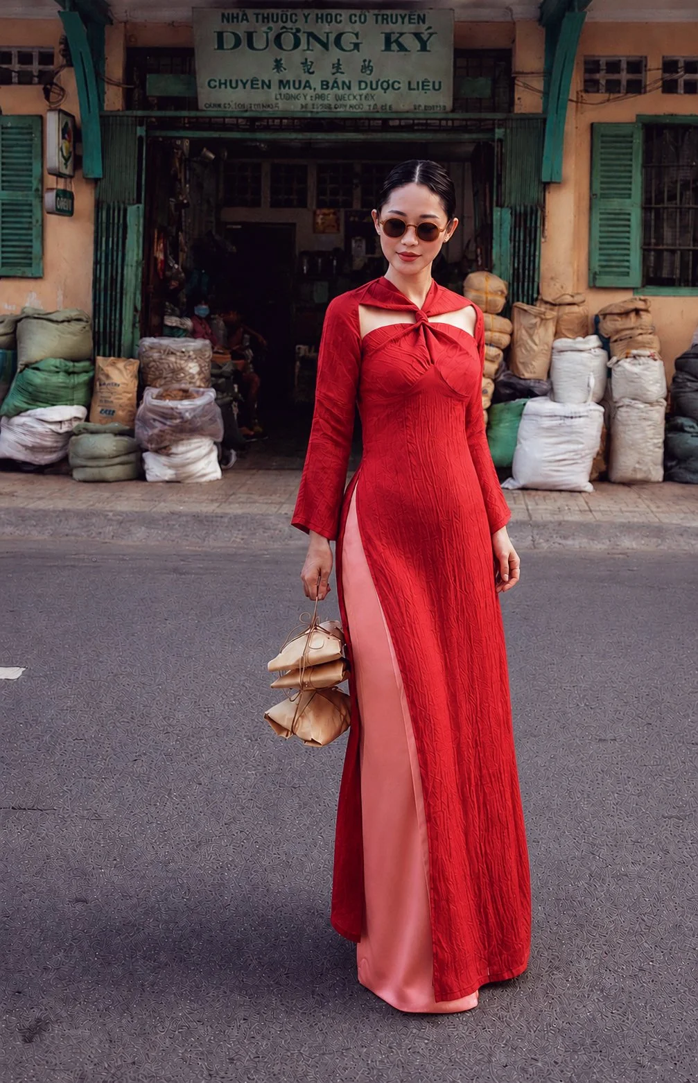 A woman wearing a red traditional dress with a long slit, standing on a street with bags of goods and a building behind her. She is holding a beige handbag and wearing sunglasses.