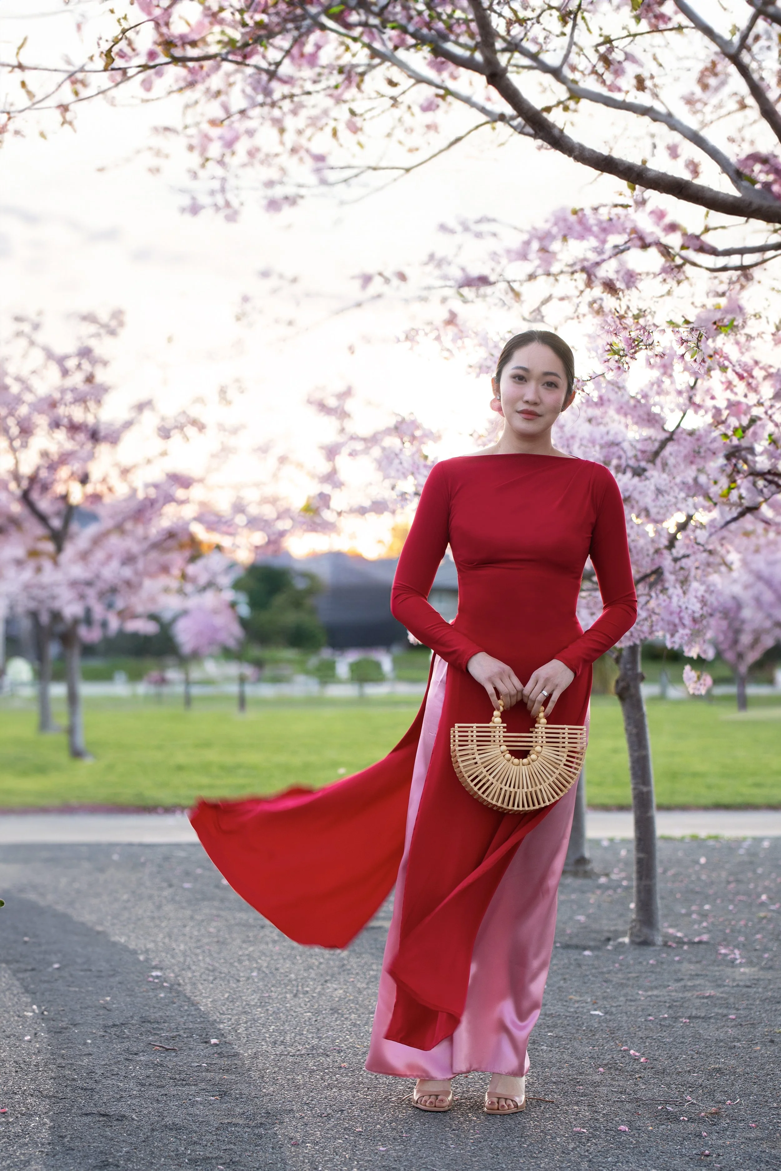 A woman in a long red dress standing outdoors on a path, holding a bamboo handbag, with pink cherry blossom trees in full bloom around her during sunset.